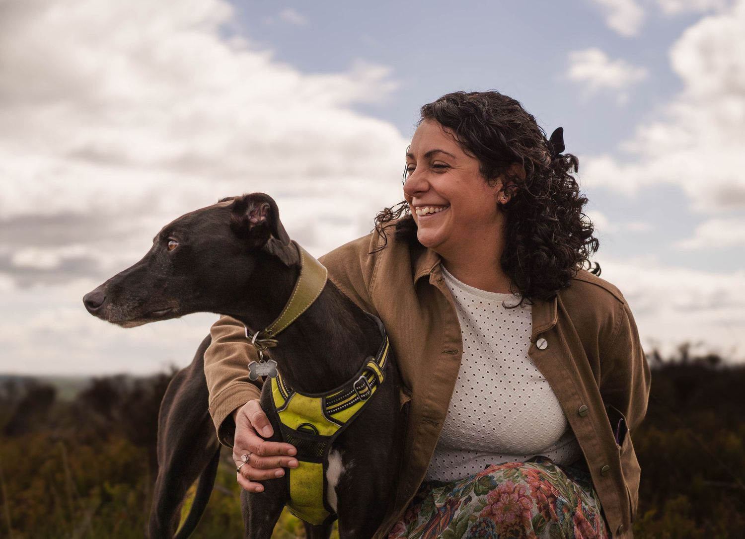 Smiling woman with curly hair sitting outdoors with a greyhound dog wearing a yellow harness.
