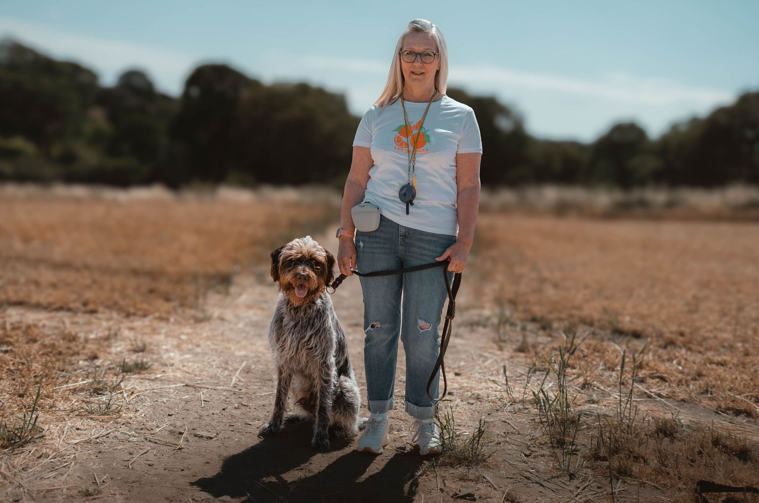 A woman stands with her small grey dog on a leash in an open sunny field.