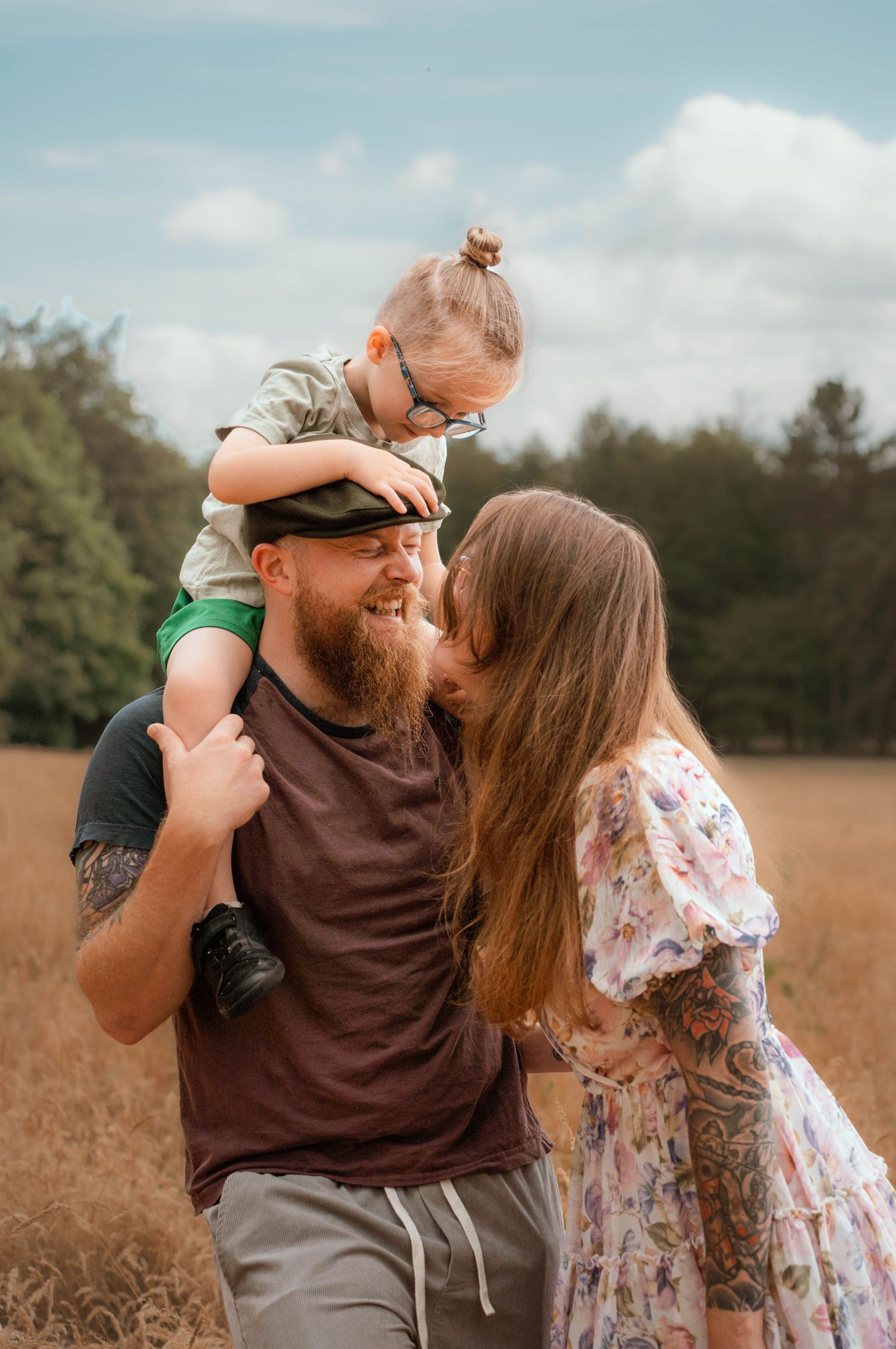 A bearded father carries his young son on shoulders while leaning in to kiss his wife in a field.