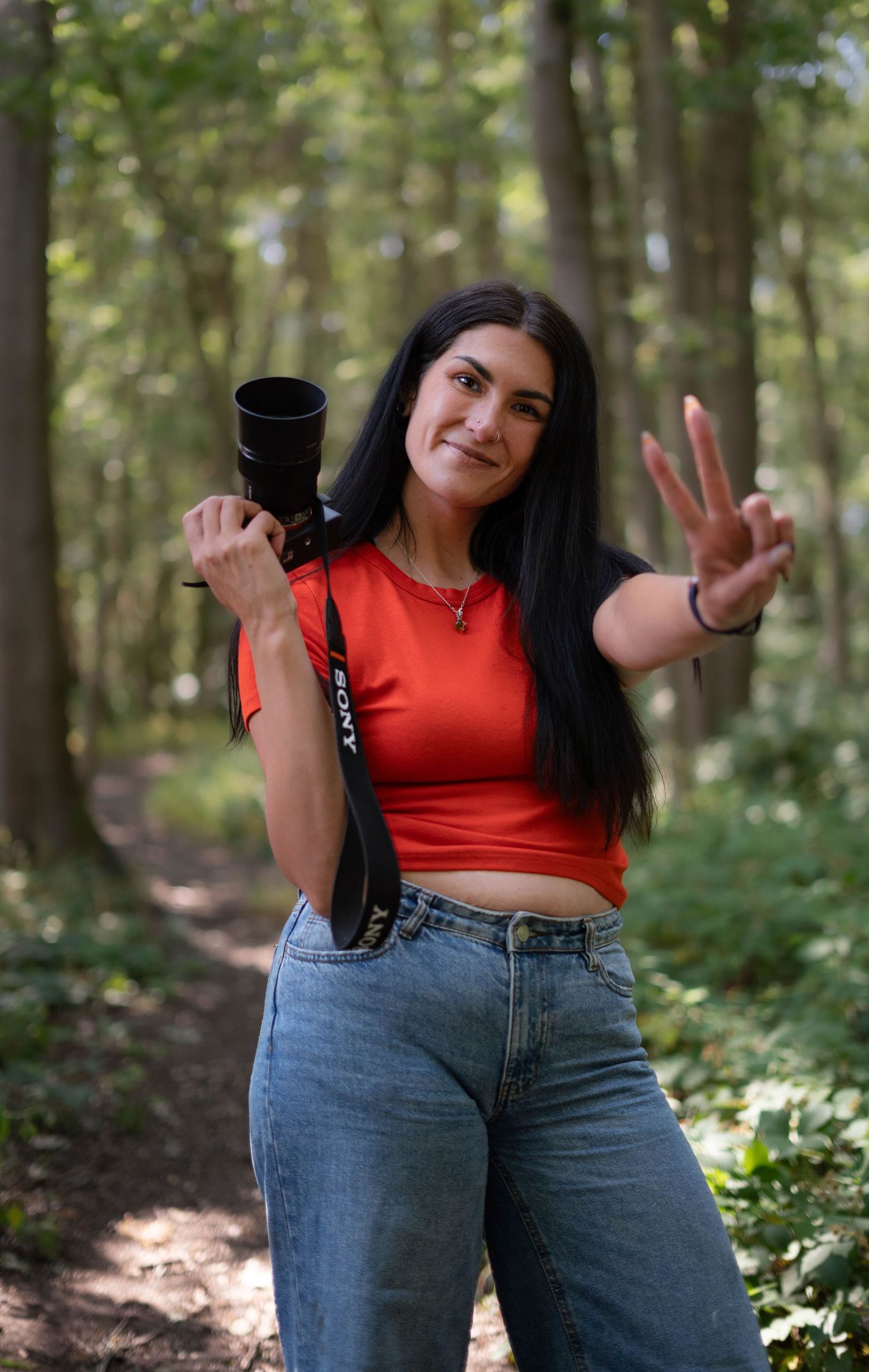 Young woman in red crop top holding a Sony camera and making a peace sign in a forest.