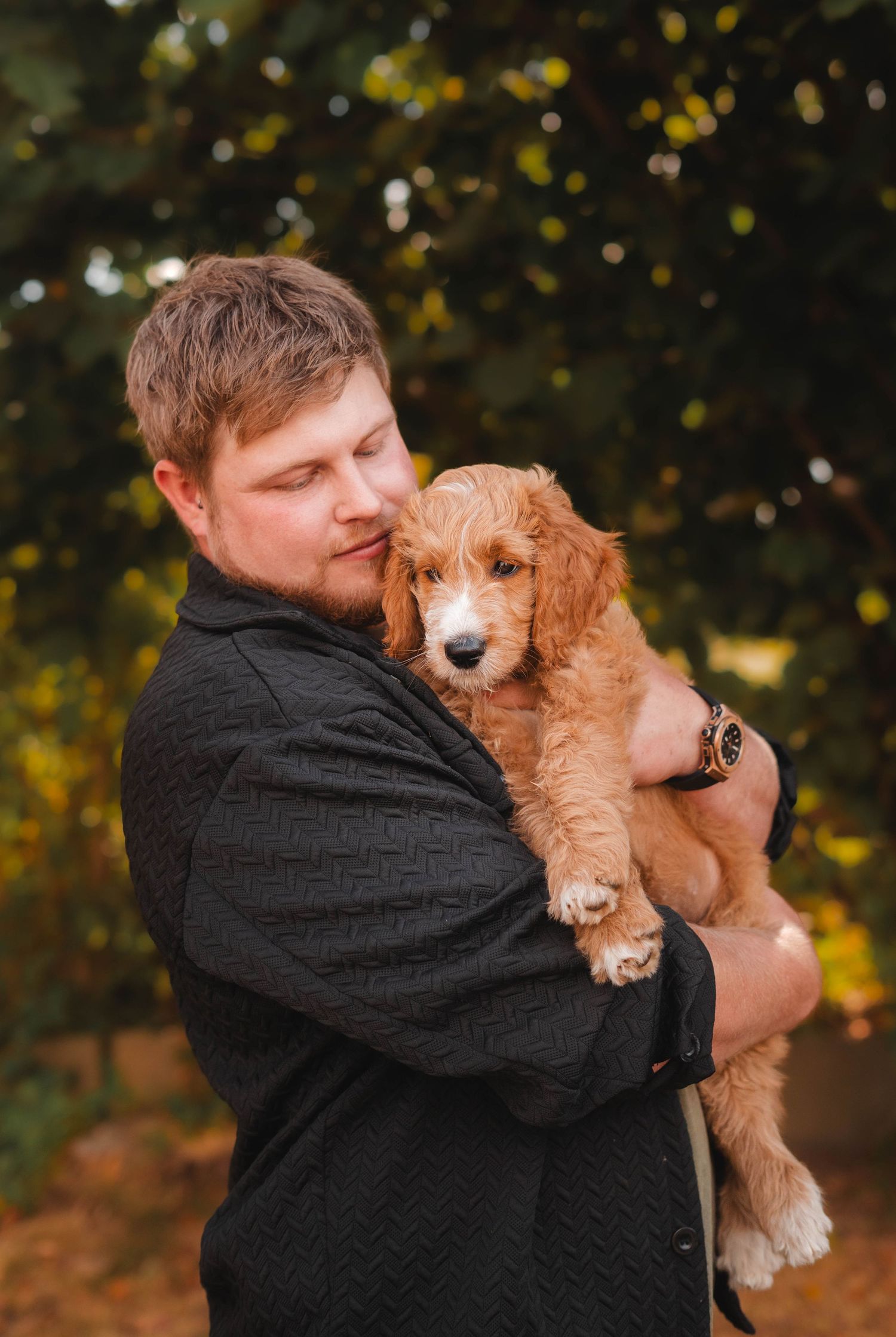 Man in black jacket tenderly holding a fluffy golden Goldendoodle puppy outdoors with green trees in background.