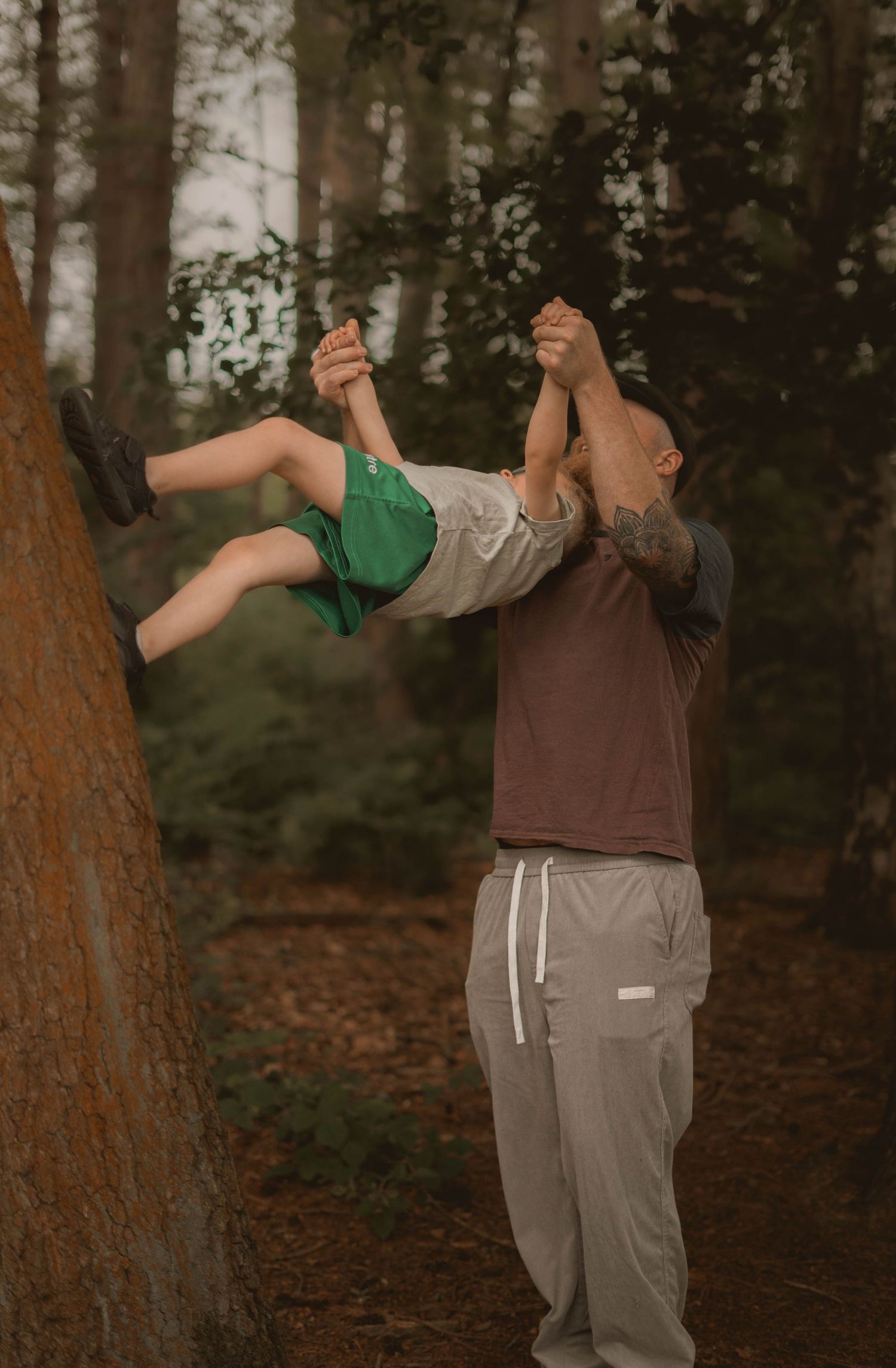 A tattooed bald father playfully lifts his child by the hands in a lush forest setting.