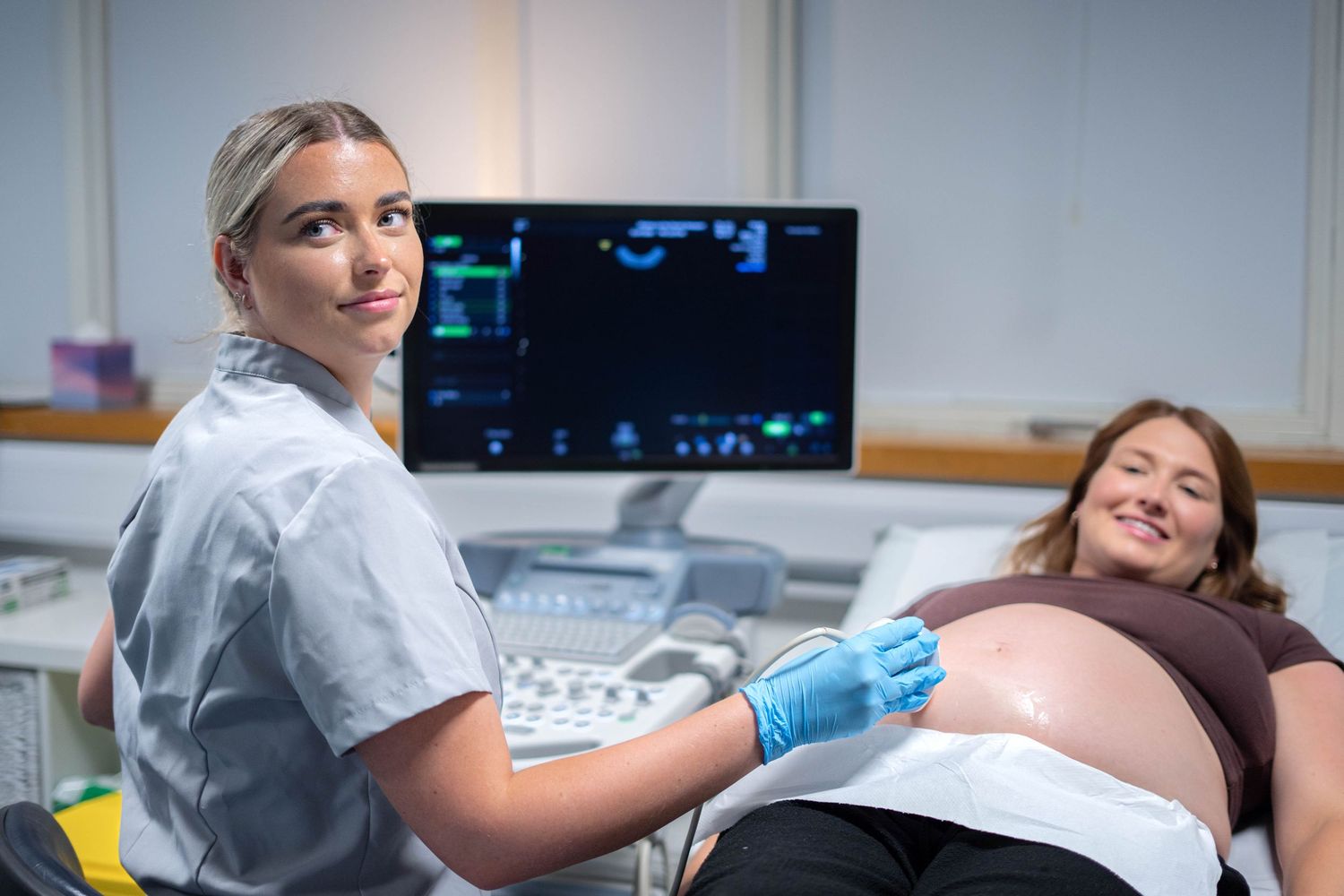 Ultrasound technician performs pregnancy scan on smiling expectant mother in clinical setting.