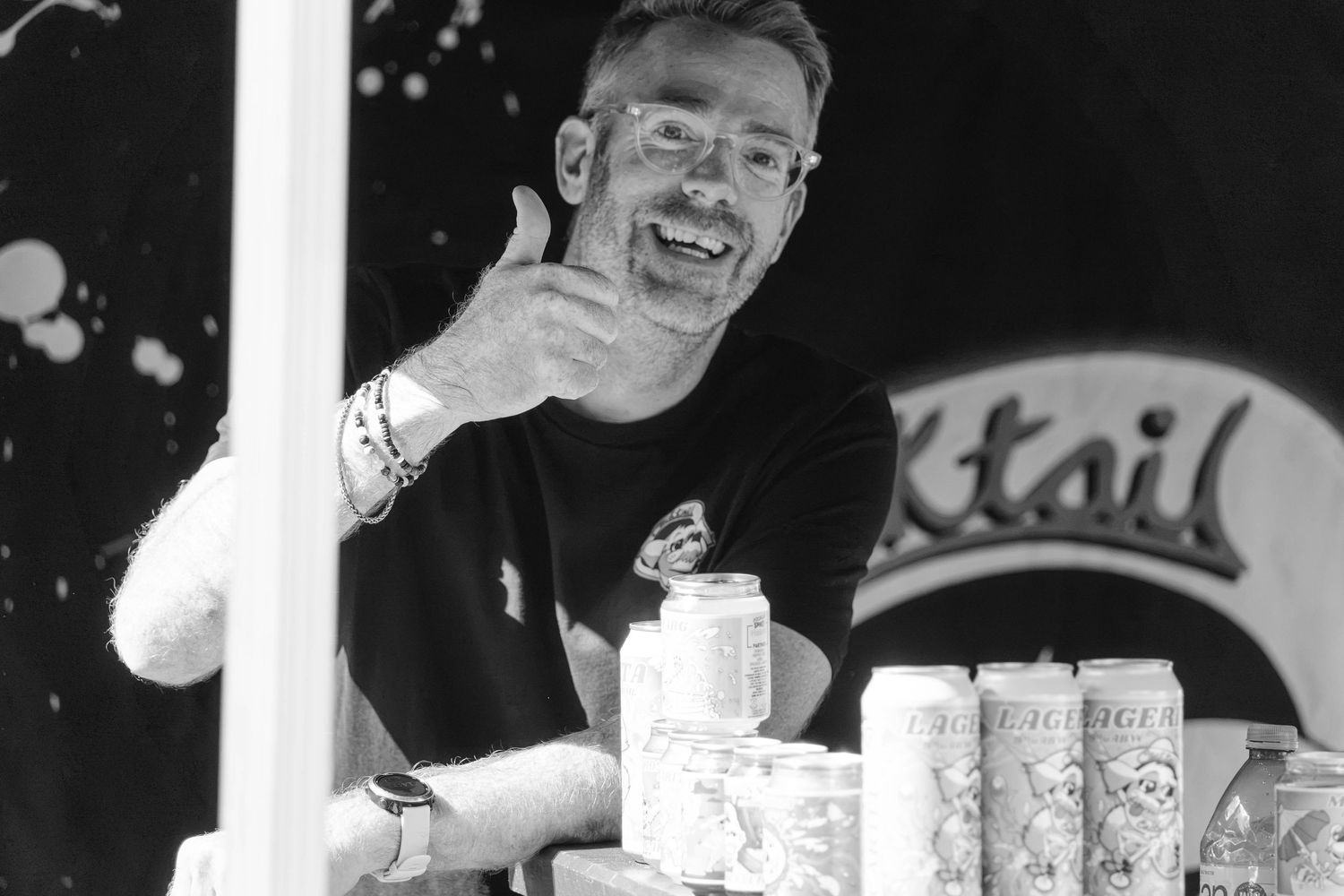 A smiling man gives a thumbs up behind a counter displaying canned beverages in black and white.