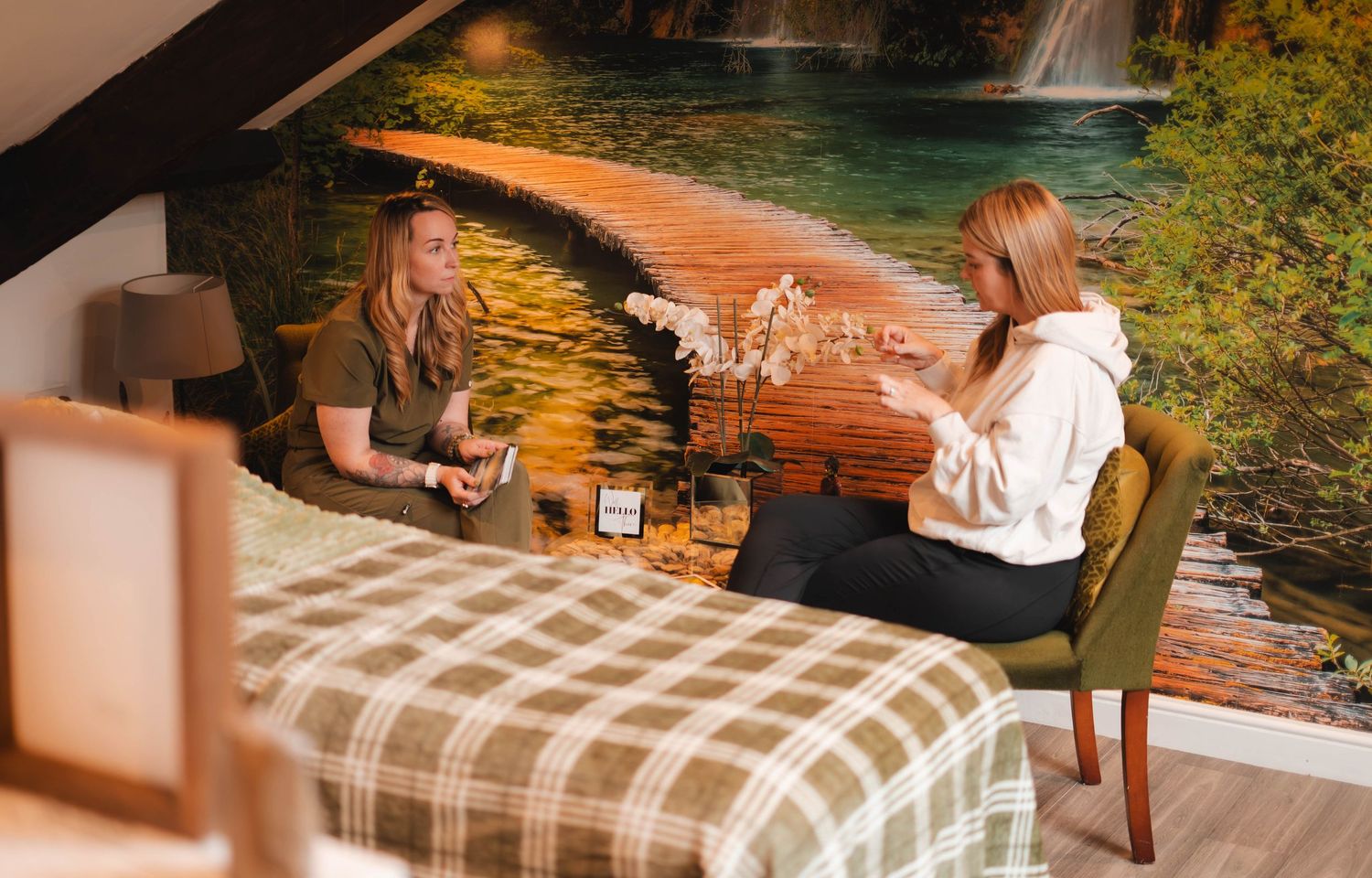Two women having a consultation session in a cozy spa room with a nature mural backdrop.