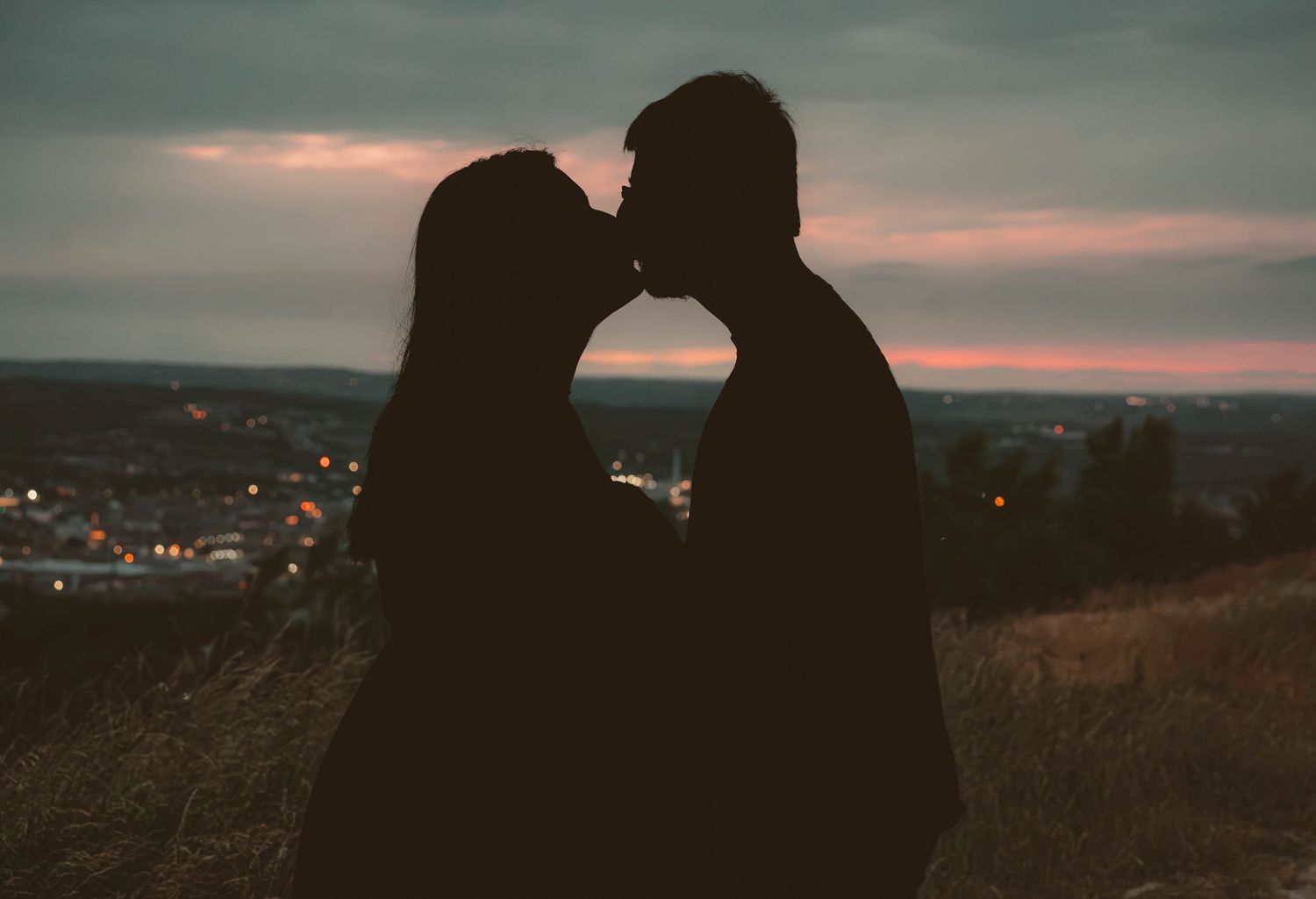 Romantic silhouette of couple kissing at dusk with city lights glowing in background.