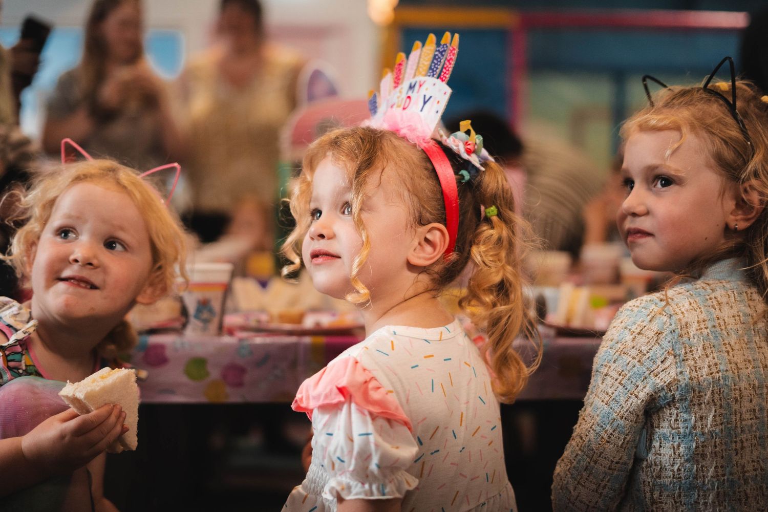 Three young girls at a birthday party, one wearing a colorful birthday crown headband, smiling joyfully.