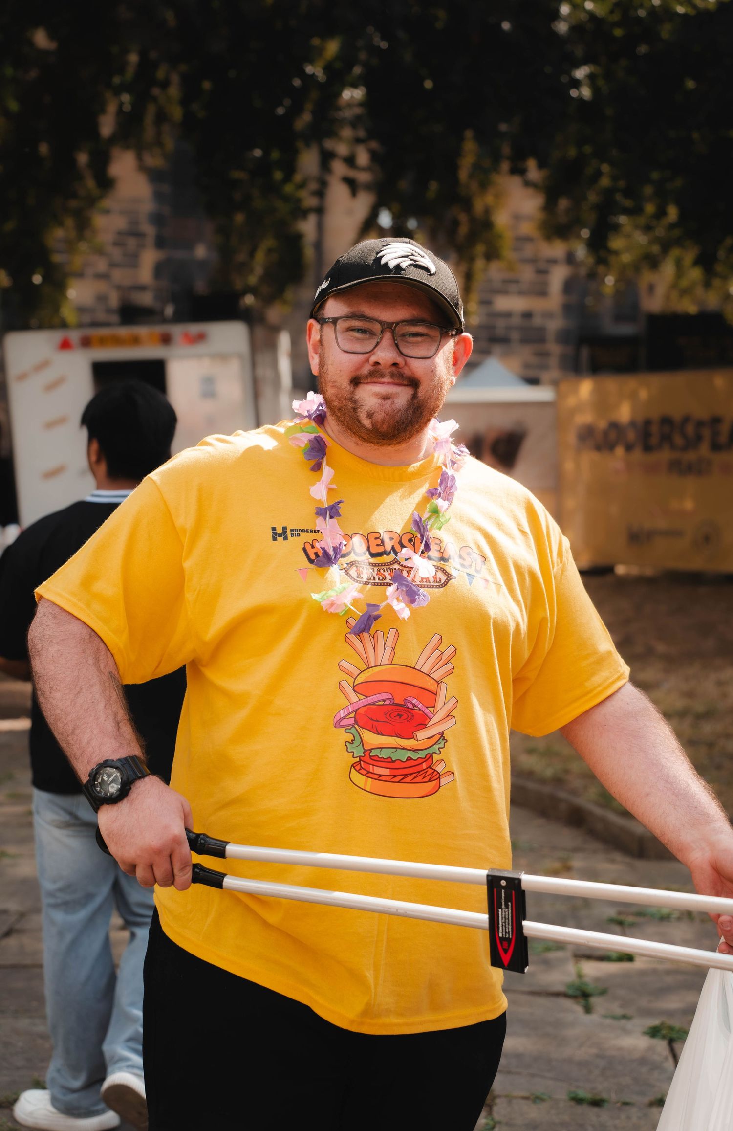 Smiling man in yellow burger t-shirt with floral lei holding crutches at outdoor event.