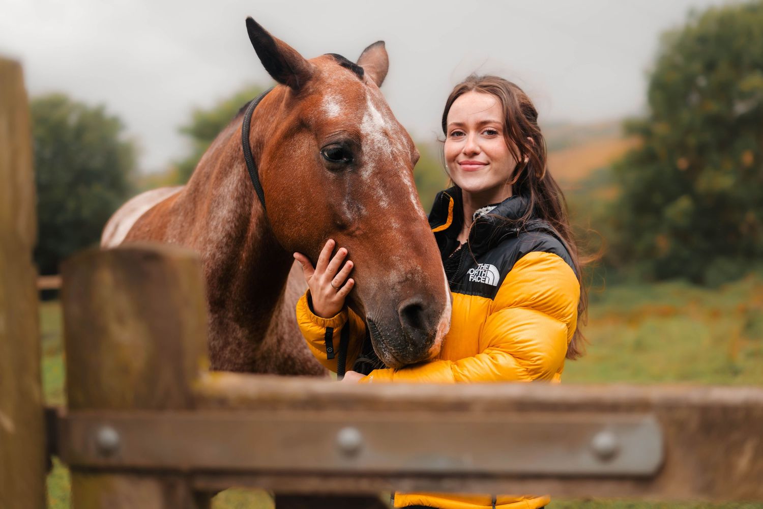 A smiling young woman in a yellow North Face jacket cuddles a chestnut horse over a wooden fence outdoors.