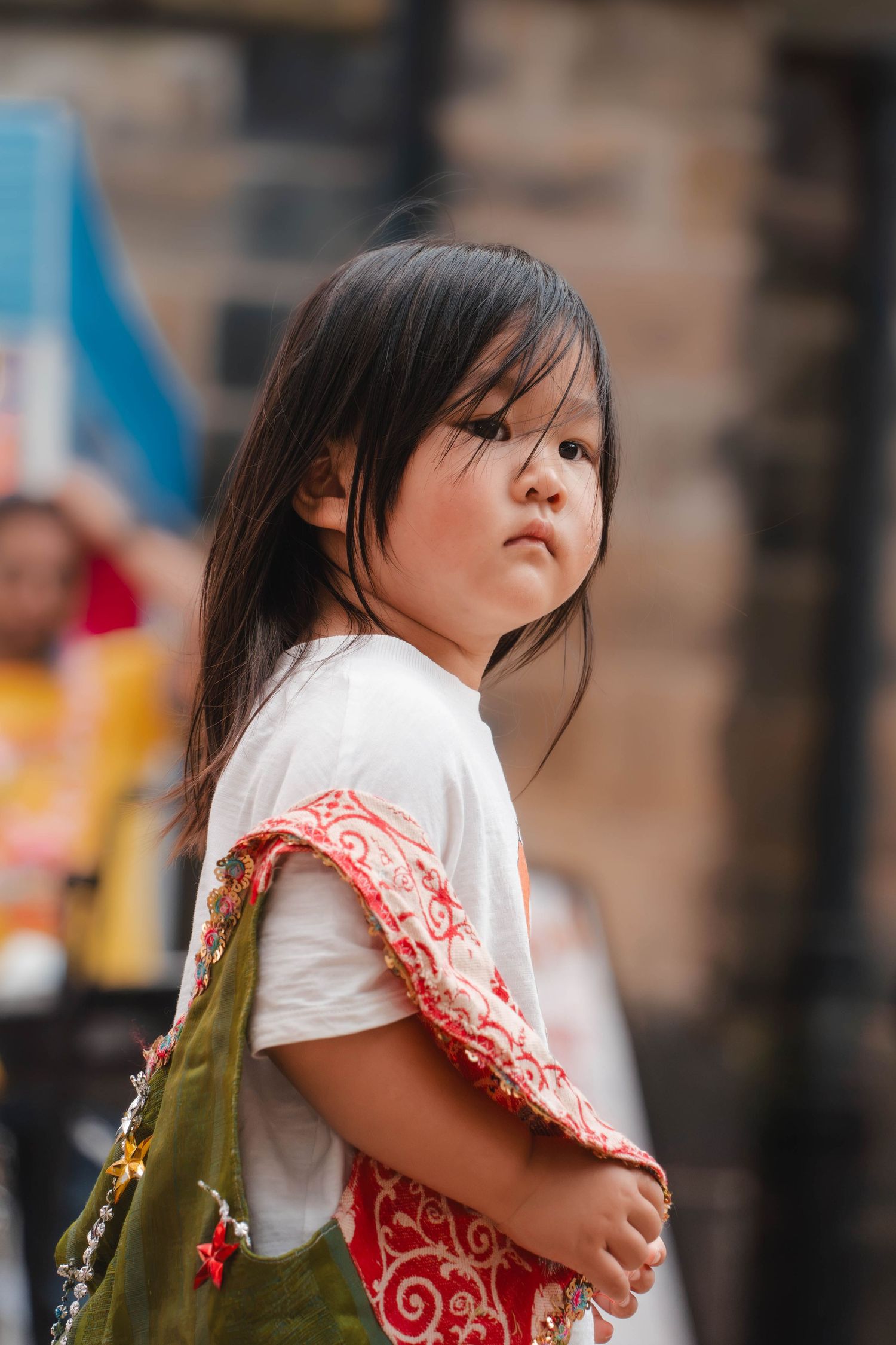 Young Asian girl with long dark hair wearing a white and red outfit carrying a green bag outdoors.