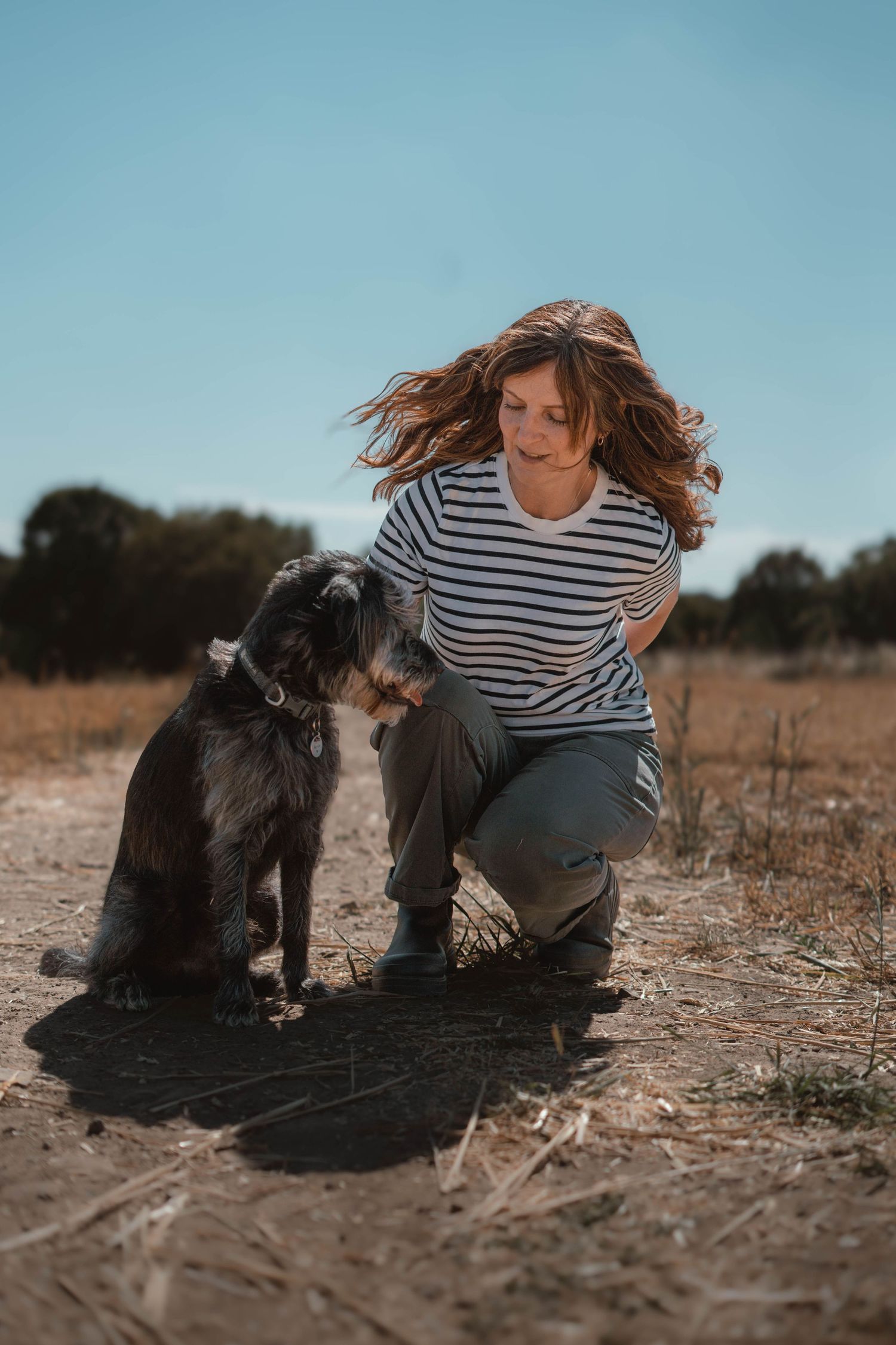A woman in a striped shirt crouches with a black and grey dog on a dirt path outdoors.