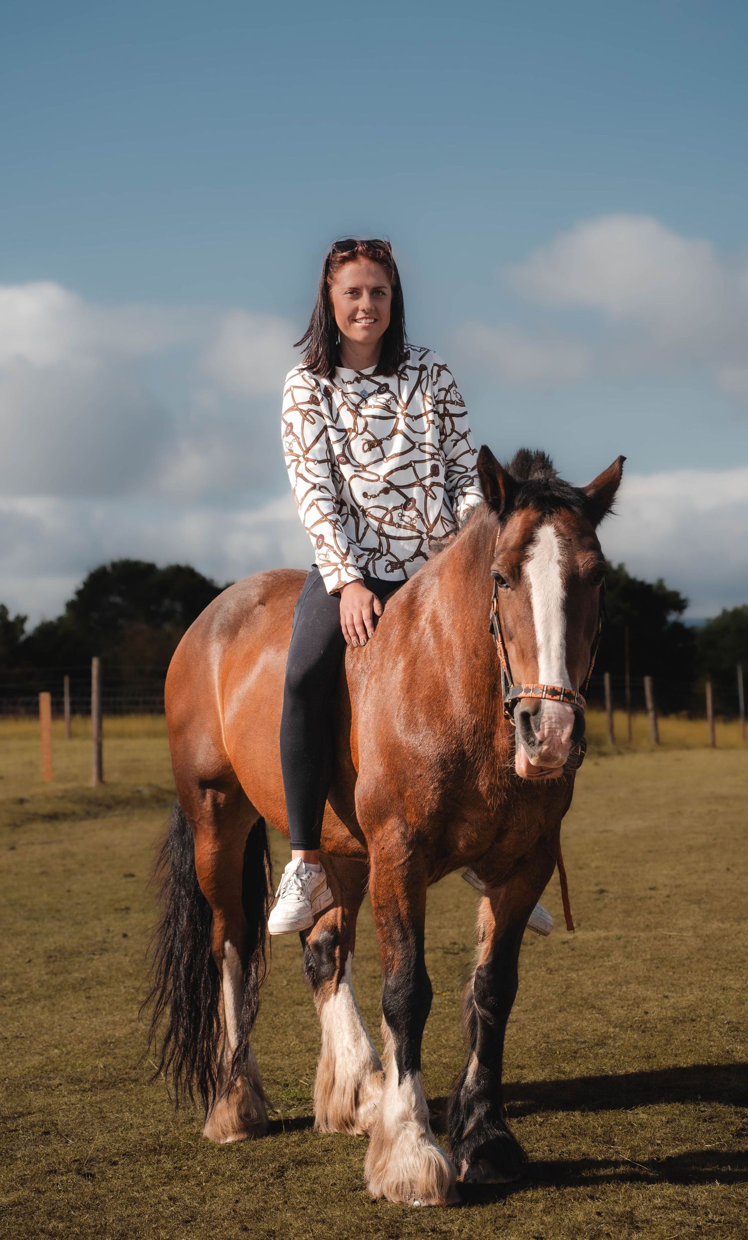 Smiling woman riding a large bay Clydesdale horse in a sunny open green field.