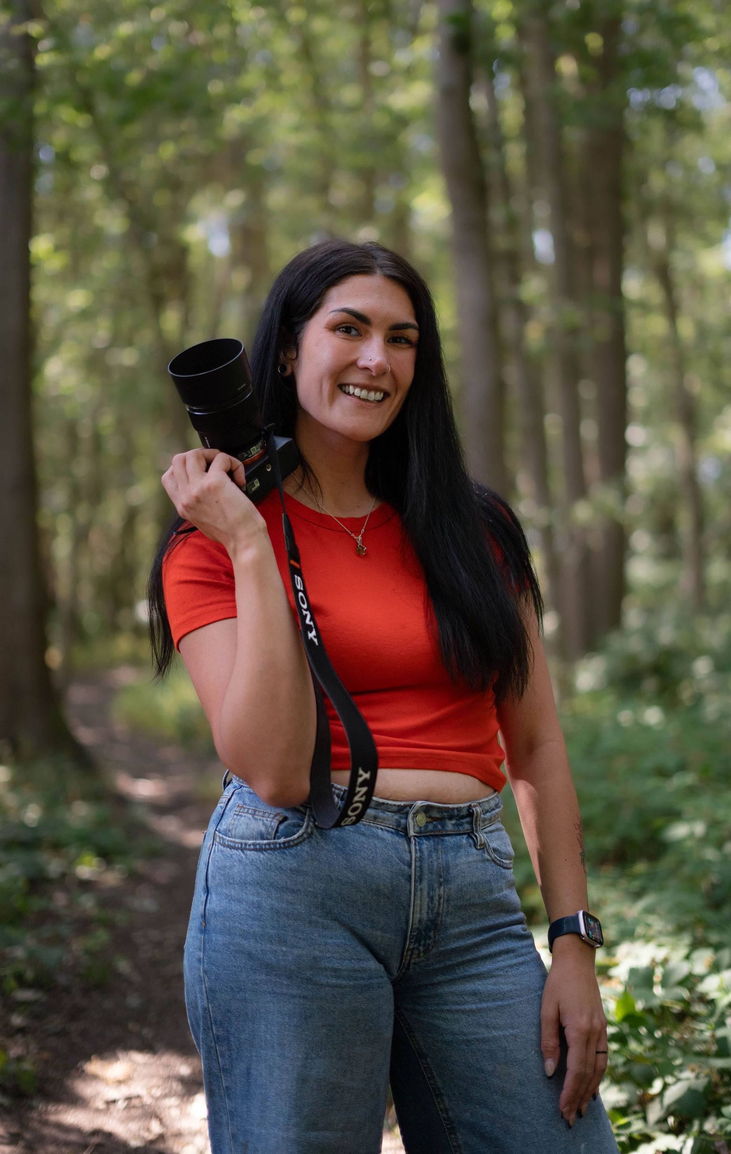 Smiling woman with long dark hair holds a Sony camera on a forest path in a red top.