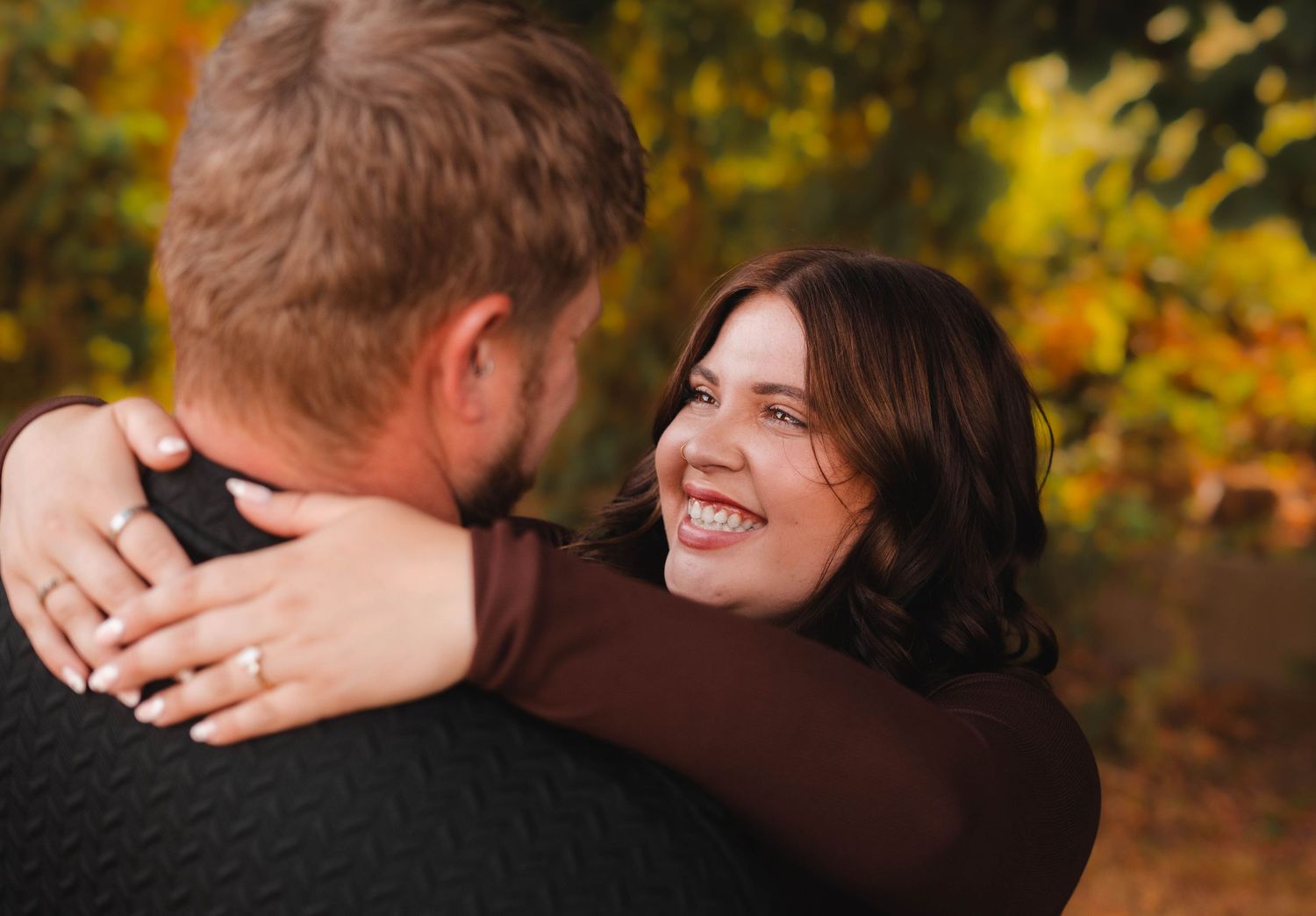 A woman with an engagement ring smiles while embracing a man outdoors in autumn.