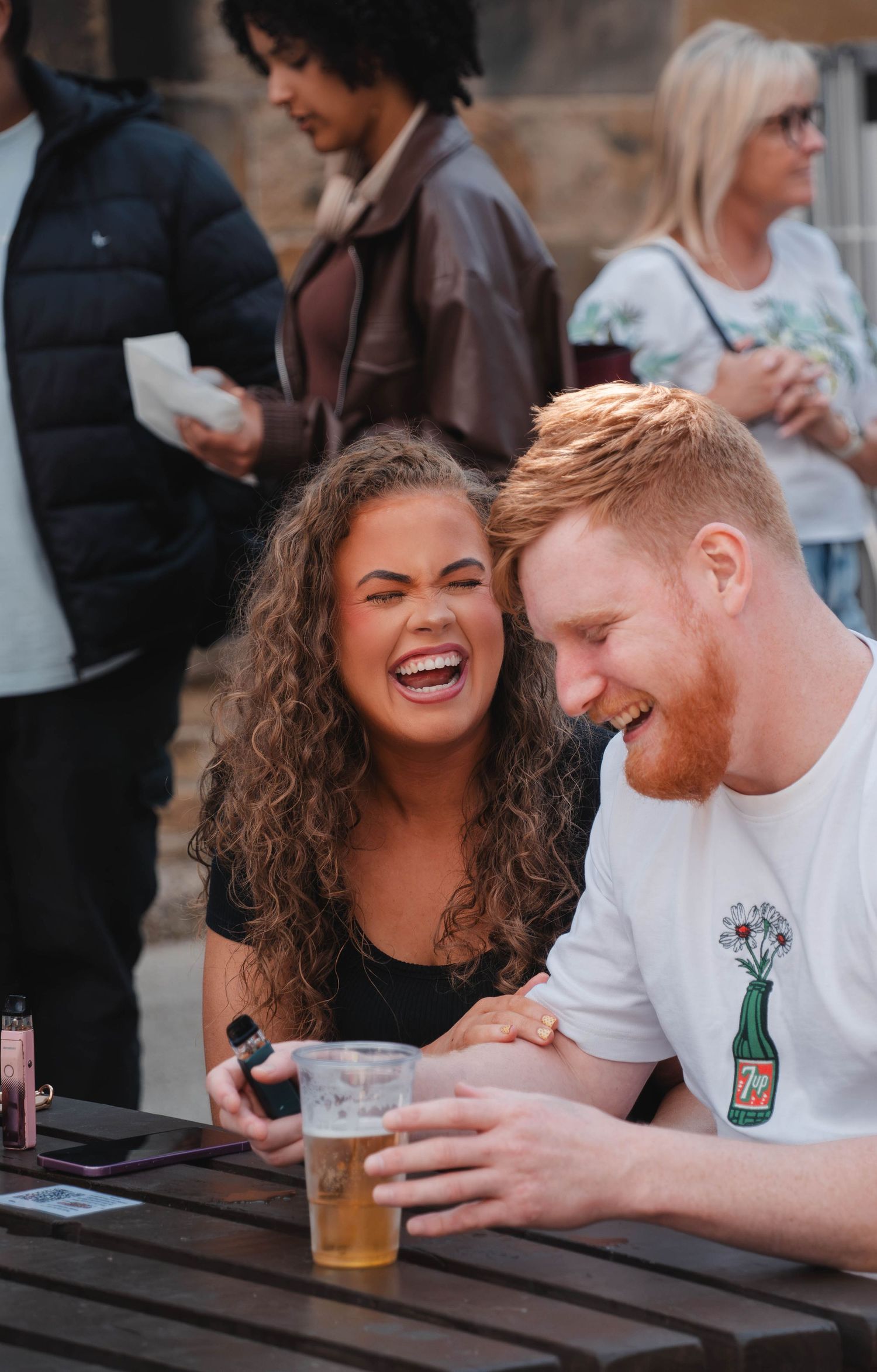 A laughing couple sits together at an outdoor table with a beer at a festival.