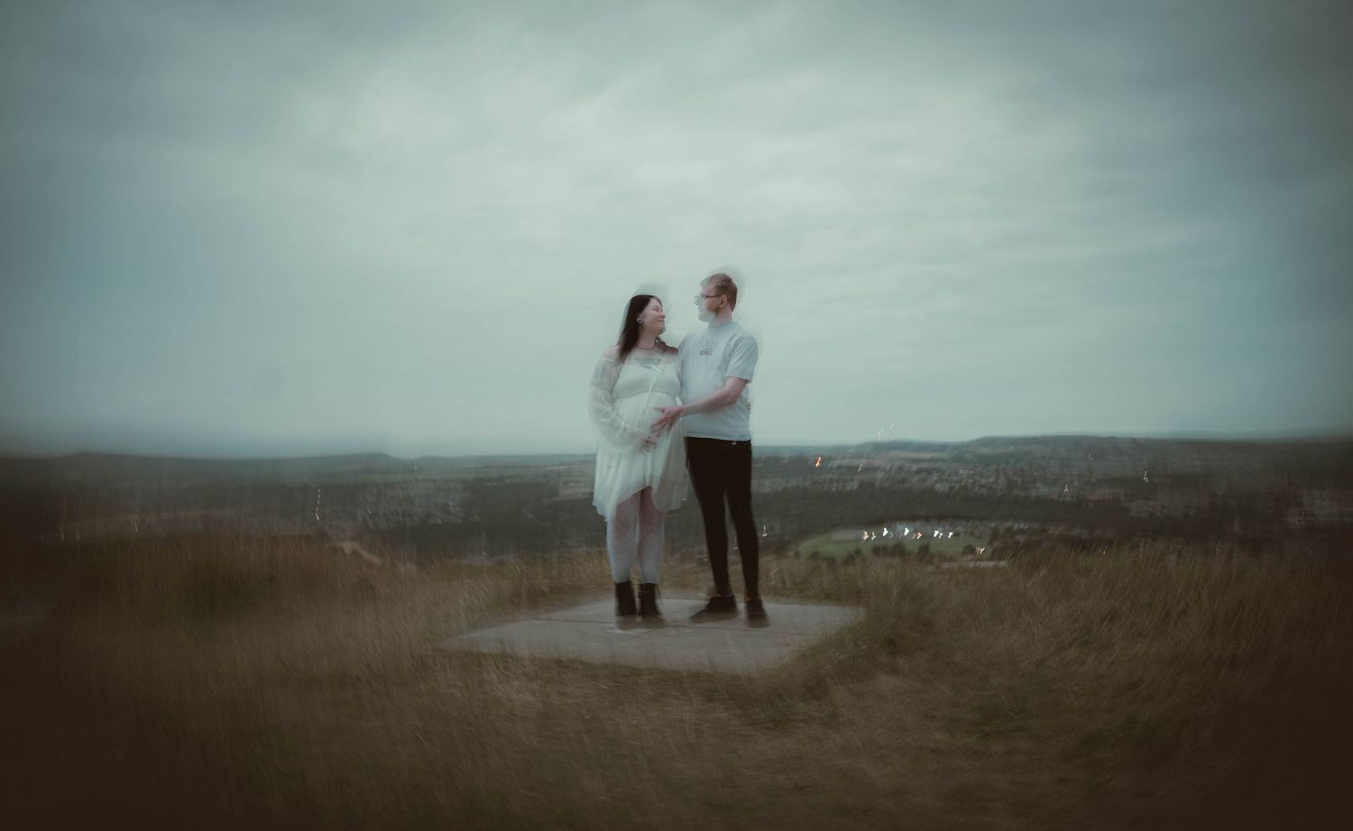 Pregnant couple standing together on hilltop overlooking town in moody overcast weather.