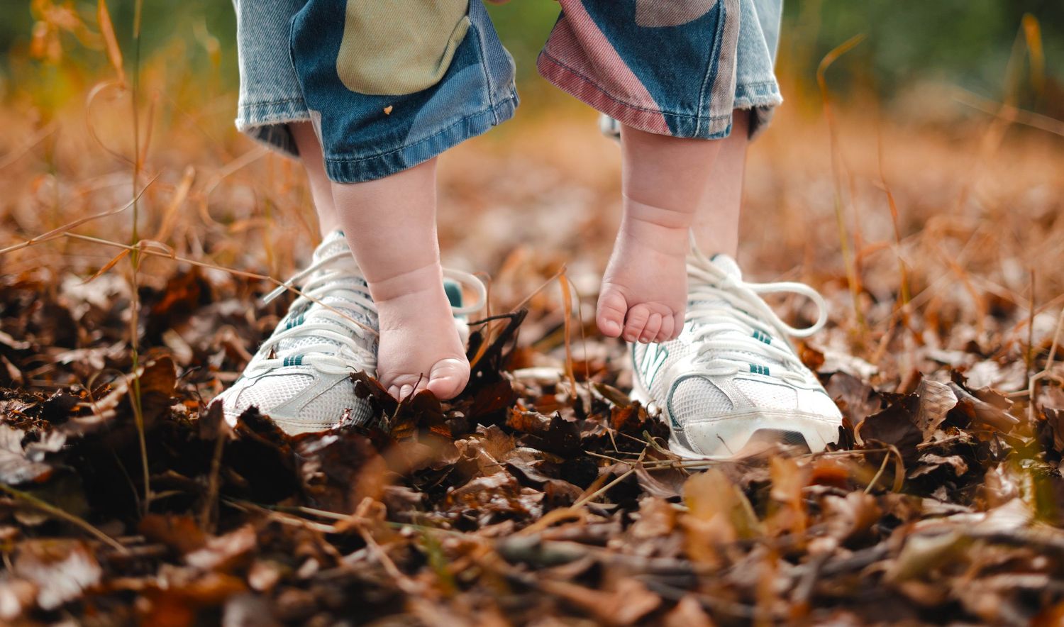 A toddler stands barefoot on an adult's sneakers among fallen autumn leaves outdoors.