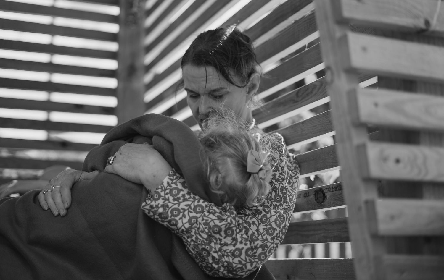 Black and white photo of woman tenderly embracing a child near wooden slat structure.