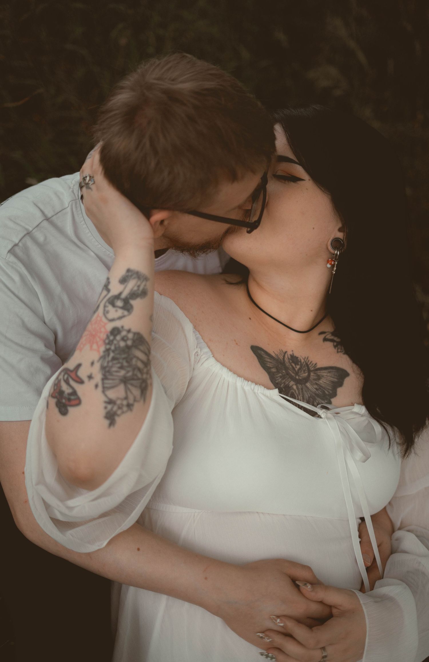 Tattooed couple sharing a kiss, woman in white maternity dress with floral arm tattoos.