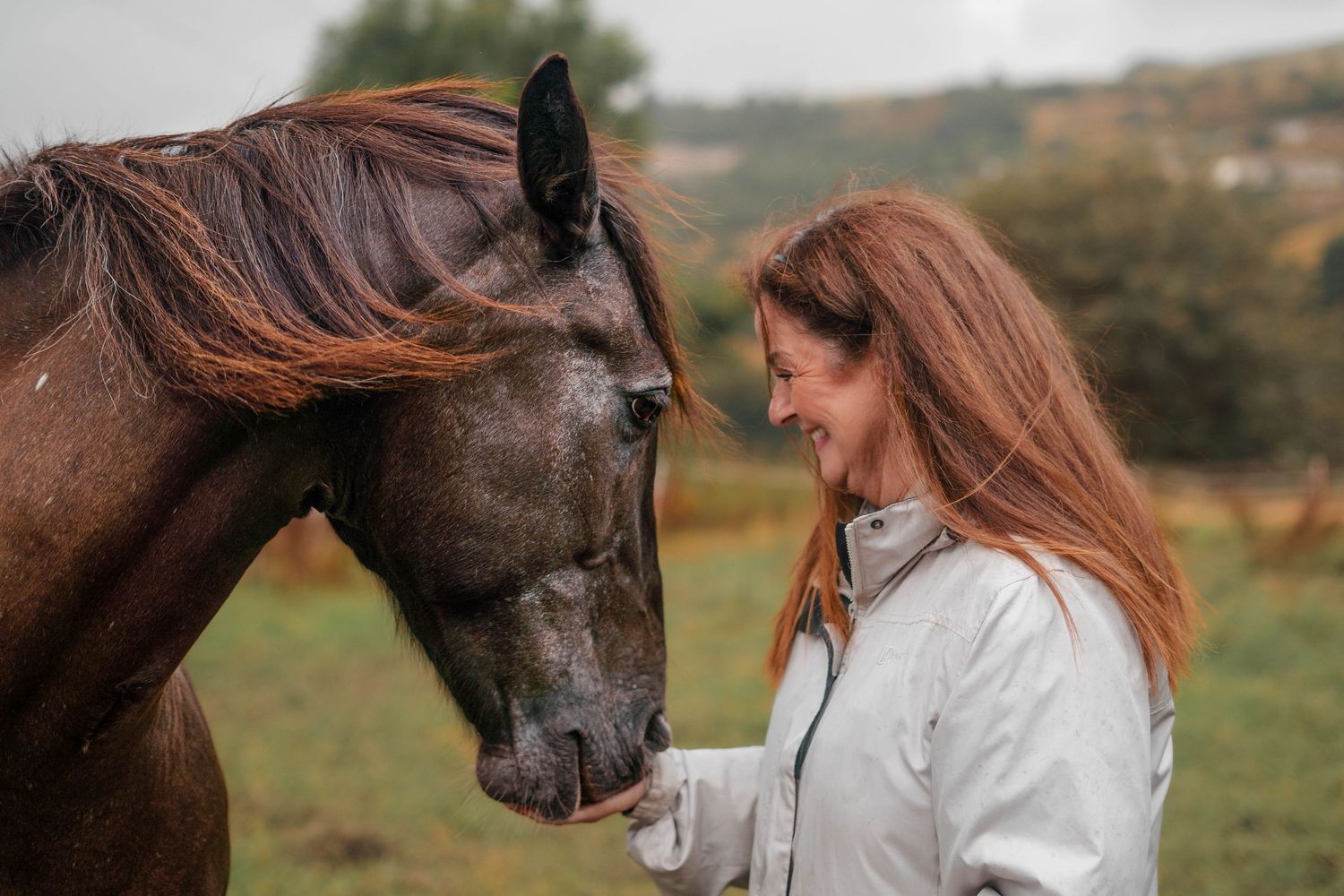 A happy redheaded woman smiles while standing close to a dark bay horse in an autumn countryside field.