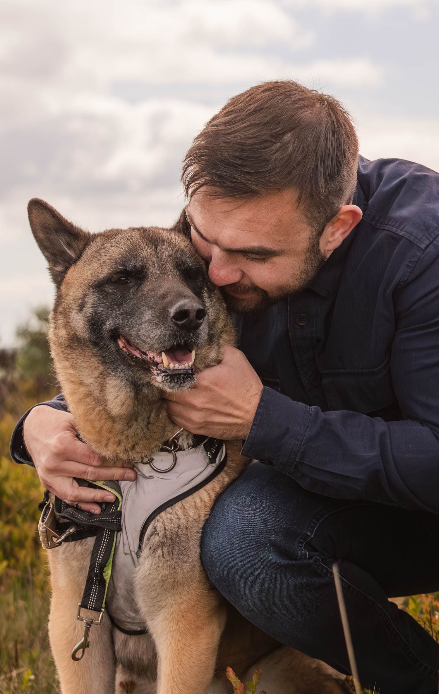 Man smiling and hugging a German Shepherd dog wearing a harness outdoors.