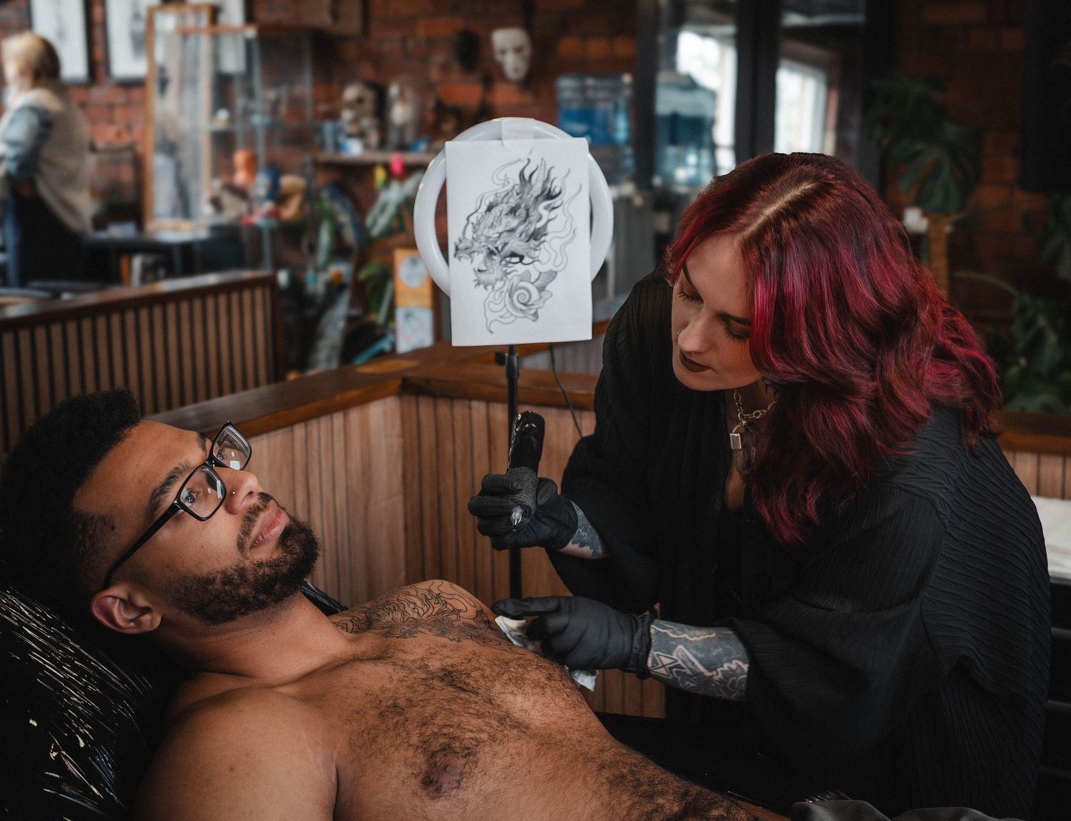 Red-haired tattoo artist carefully works on a chest tattoo design on a male client.