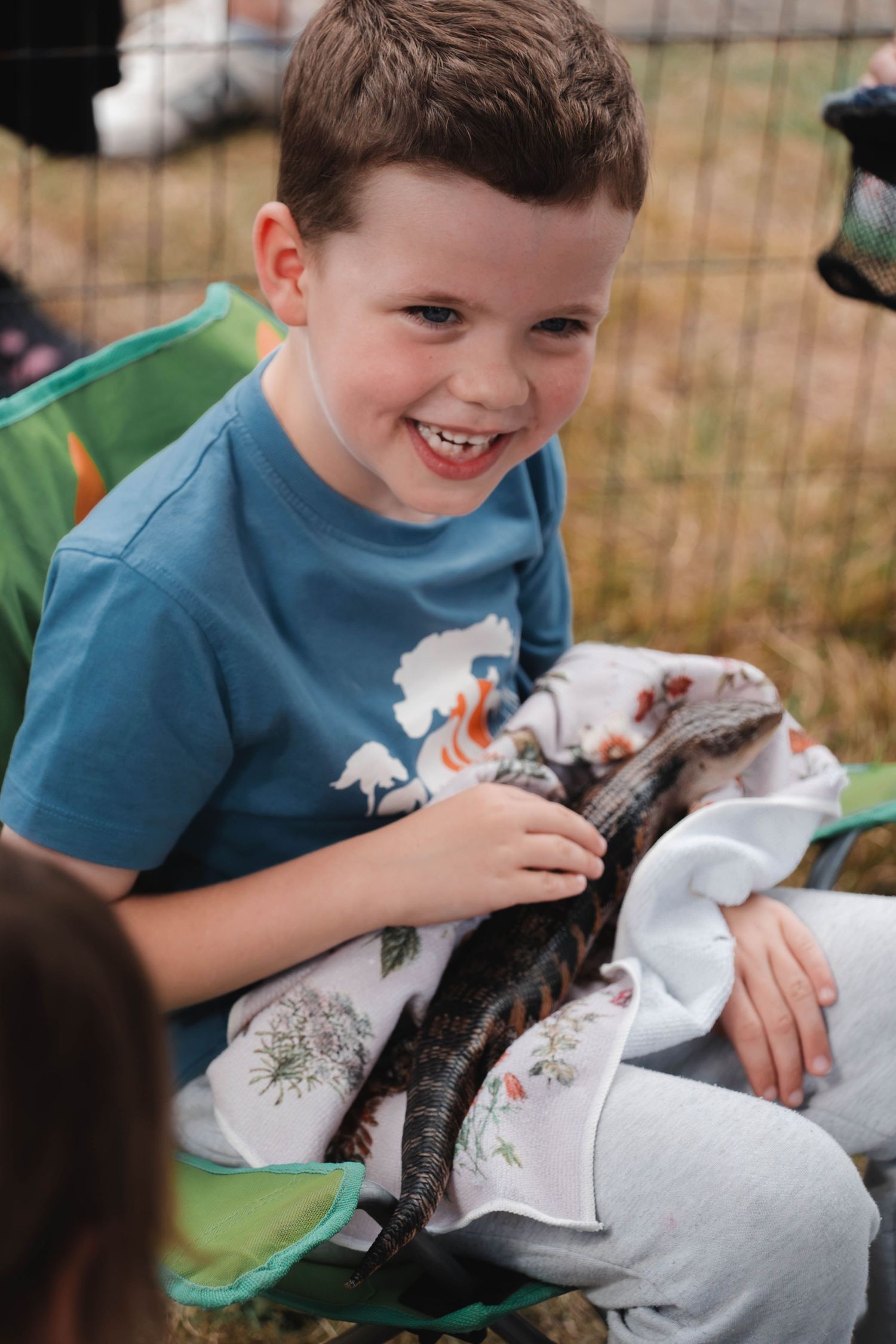 Happy young boy smiling while holding a small lizard wrapped in a blanket at an outdoor event.