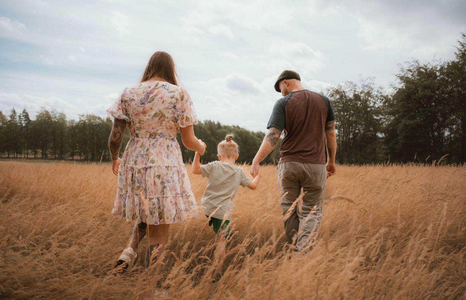 Family of three walking hand in hand through a golden wheat field under cloudy skies.