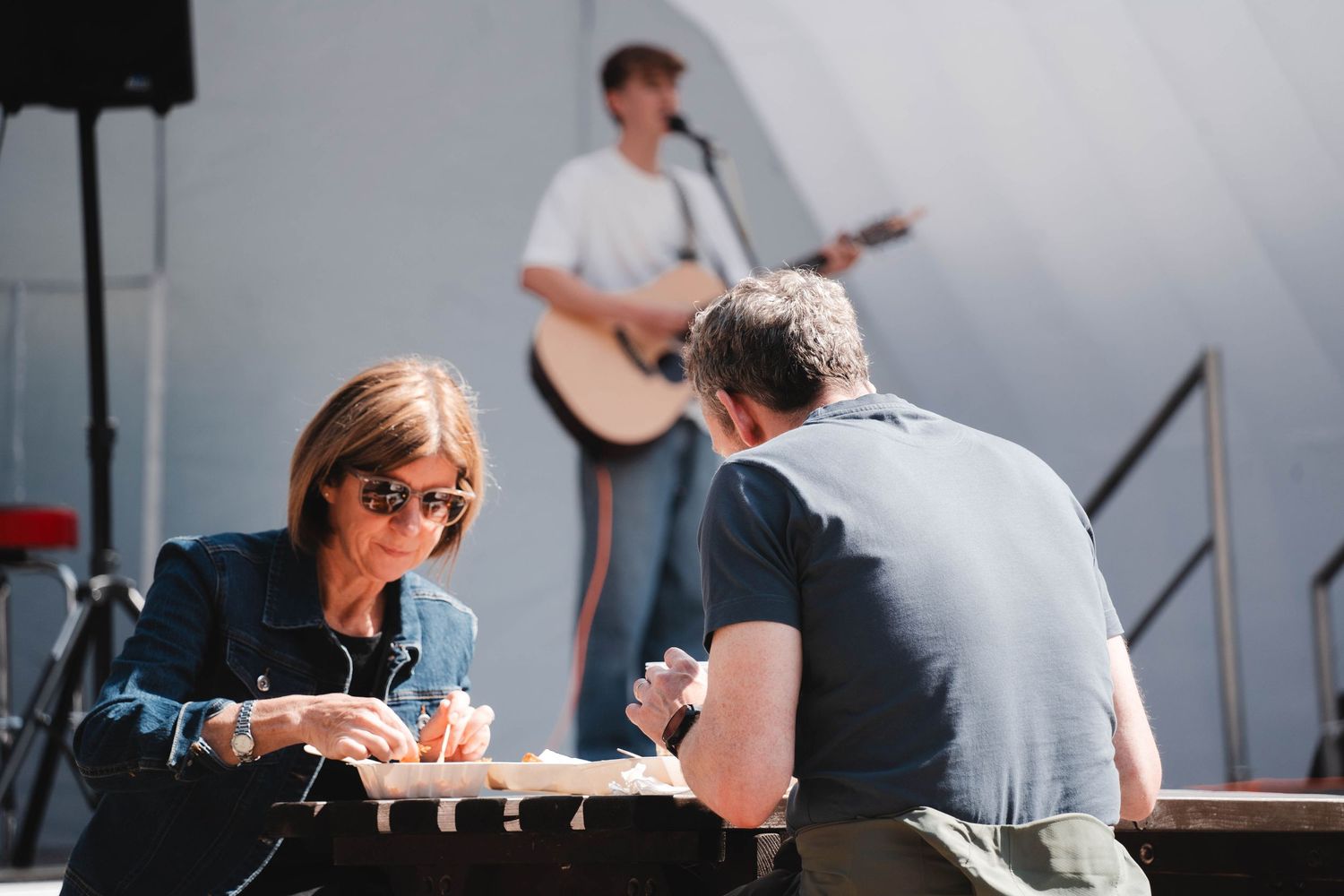 Two people engaged in conversation at an outdoor event with a guitarist performing on stage.