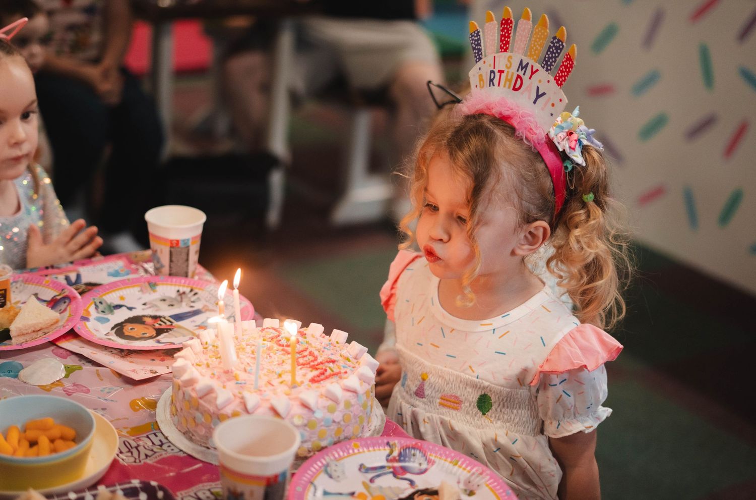 Young girl in birthday crown blowing out candles on pink birthday cake at party table.