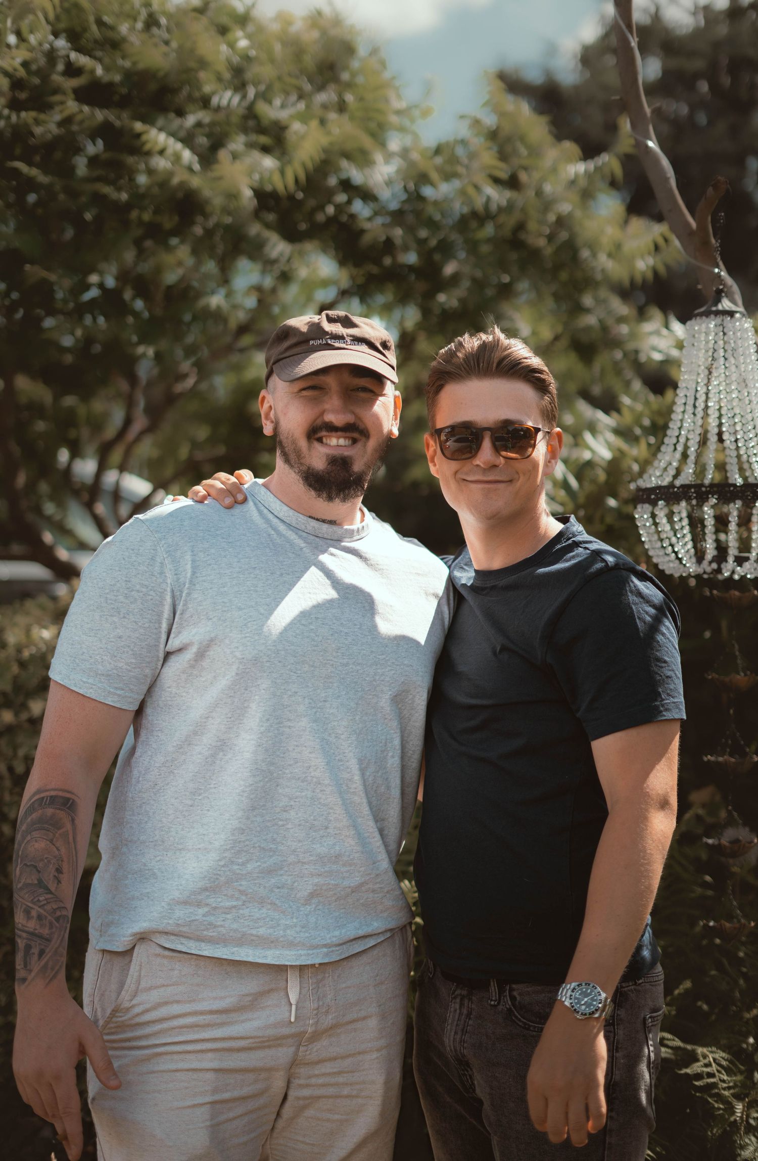 Two smiling men posing together outdoors surrounded by lush green trees on a sunny day.