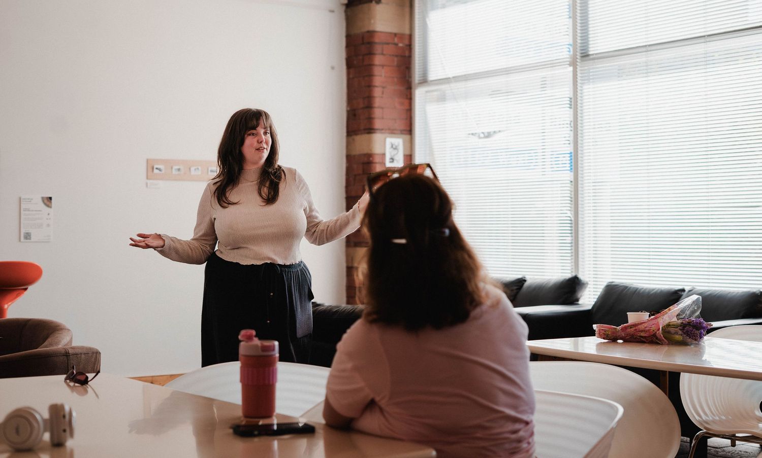 Woman presenting and gesturing while speaking to a seated person in a modern office space.