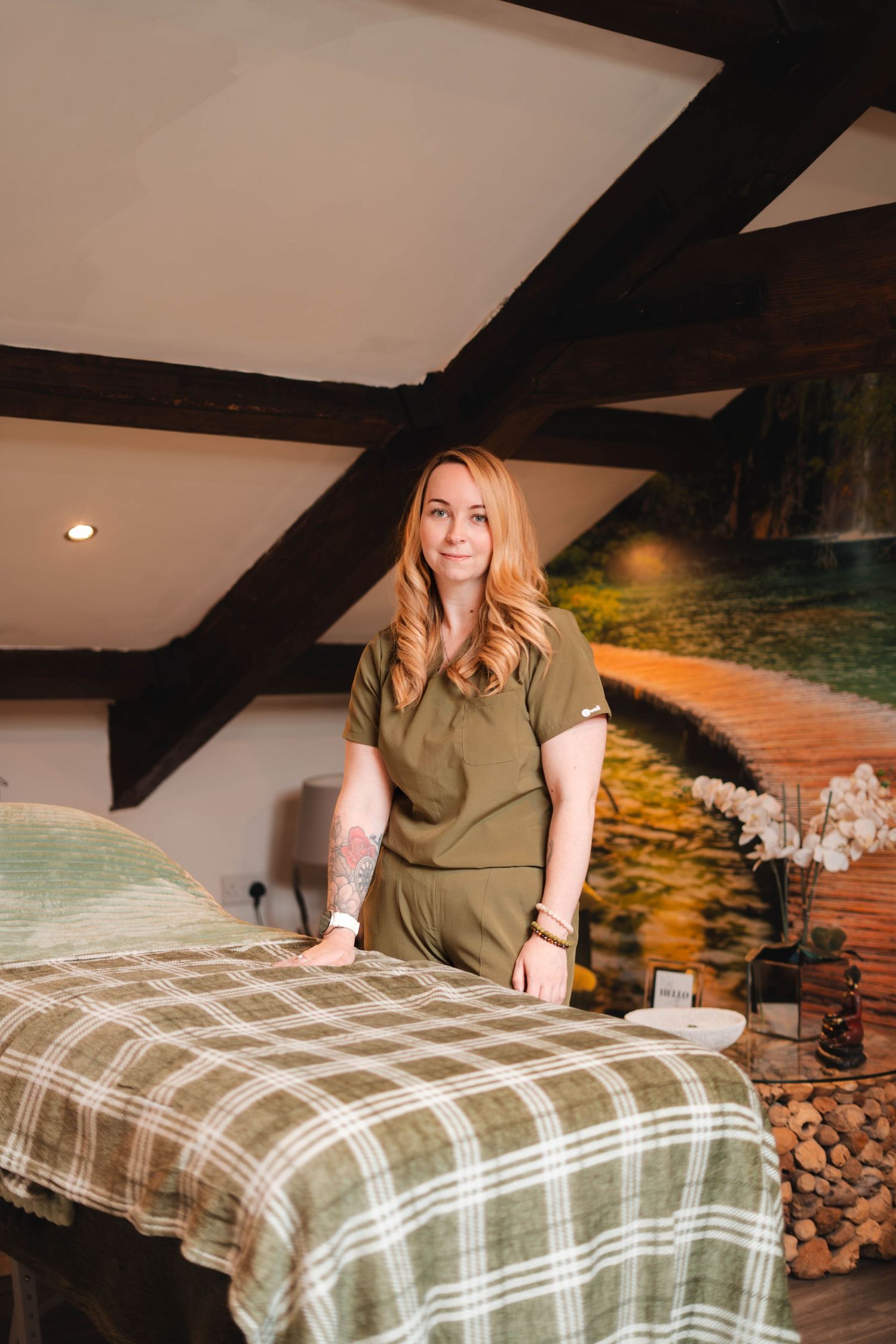 Female therapist in olive scrubs standing beside massage table in cozy spa treatment room.