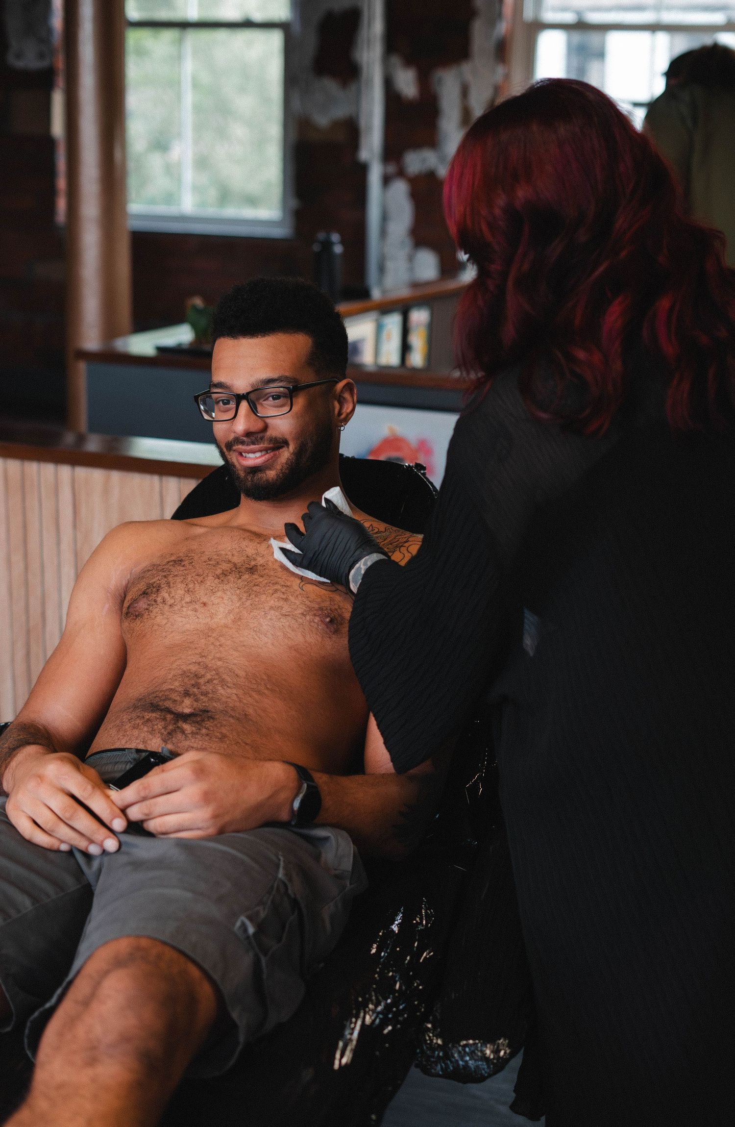 Female tattoo artist with red hair prepares to tattoo a smiling shirtless male client.