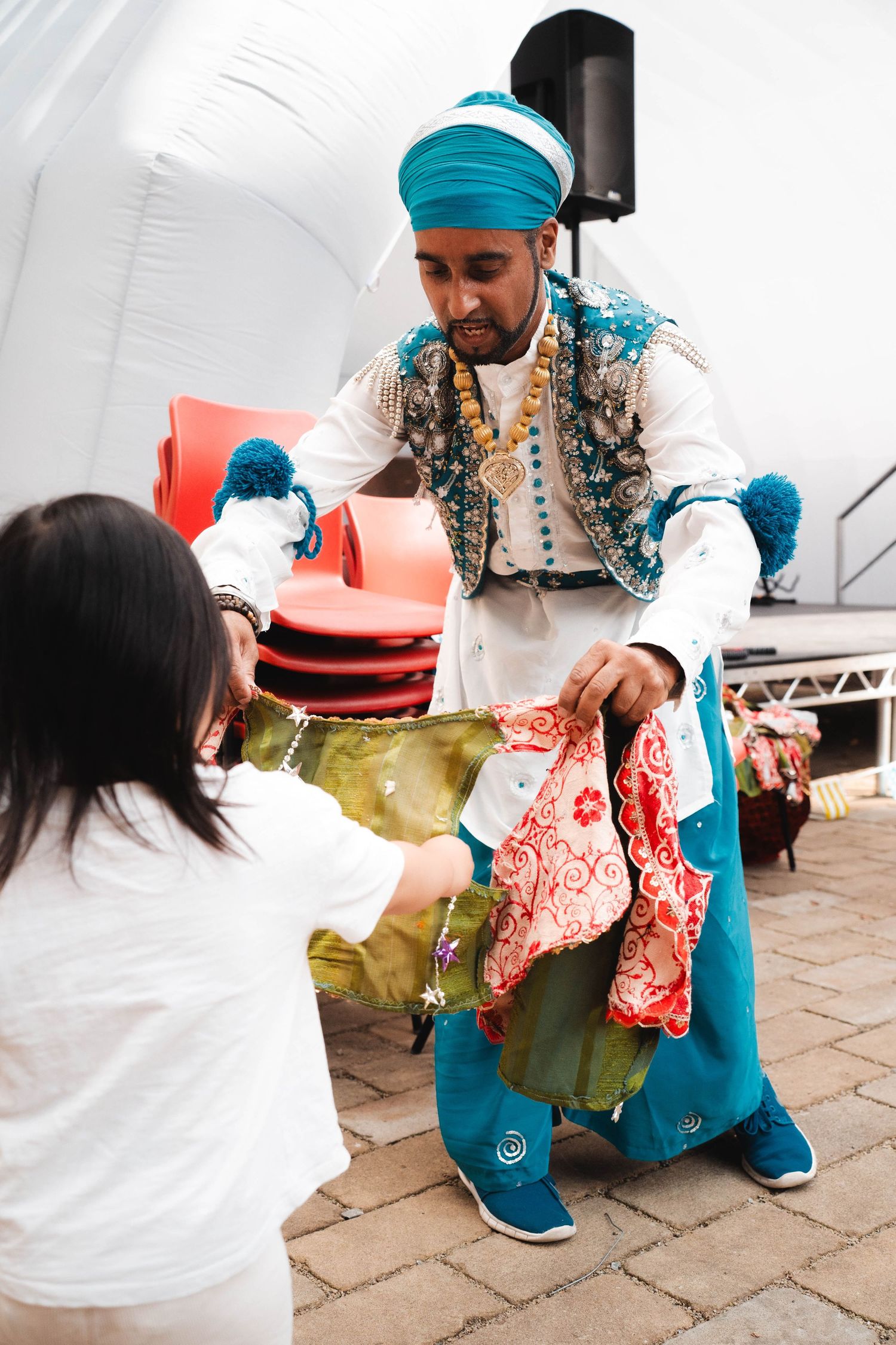 Performer in traditional Indian turban and costume handing colorful fabric to child on stage.