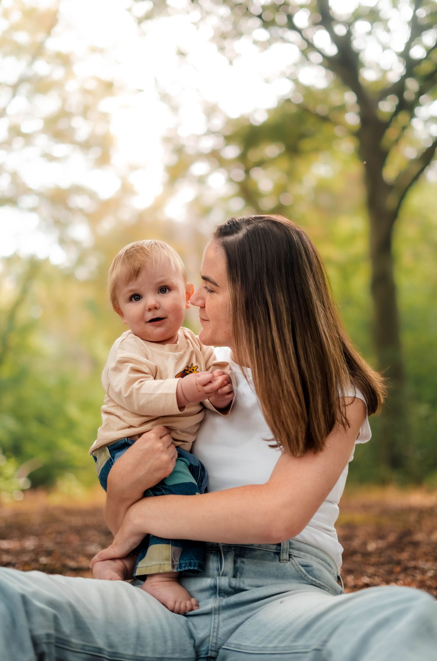 Young mother sitting outdoors in a sunlit park holding her adorable baby boy in autumn leaves.