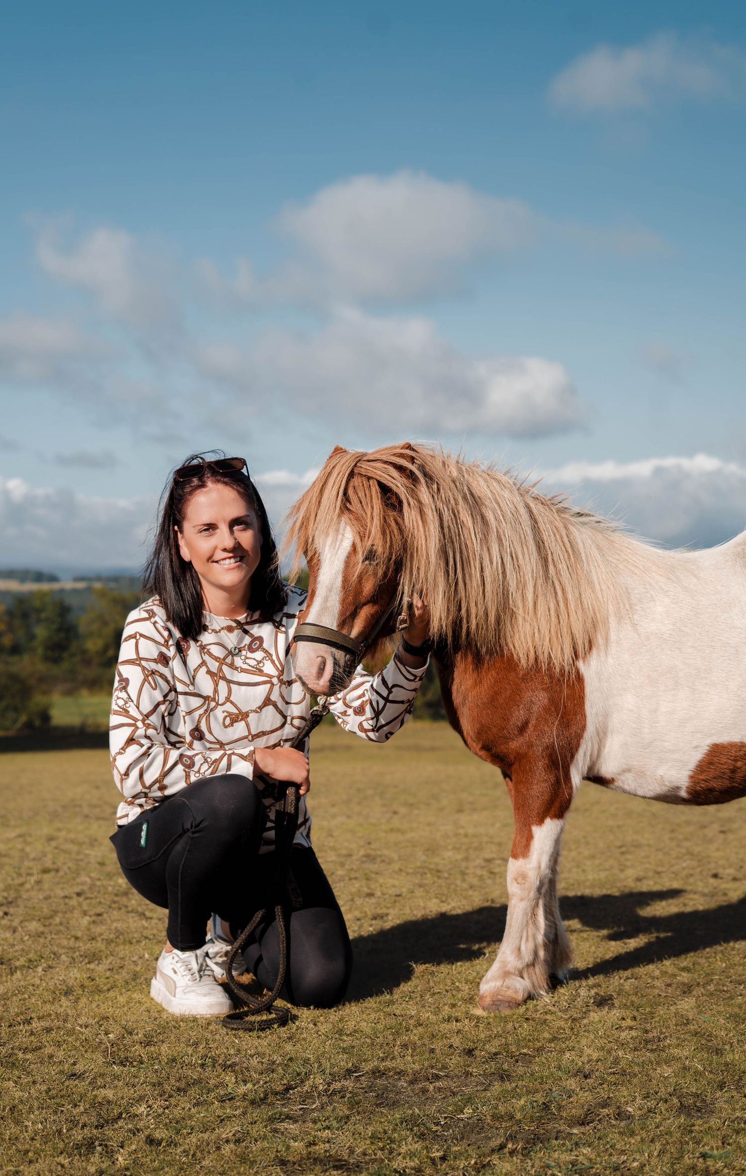 Smiling woman crouching beside a brown and white Shetland pony in a sunny open field.