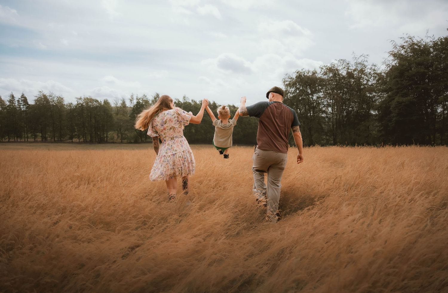 Family of three joyfully running through a golden field holding hands on a cloudy day.