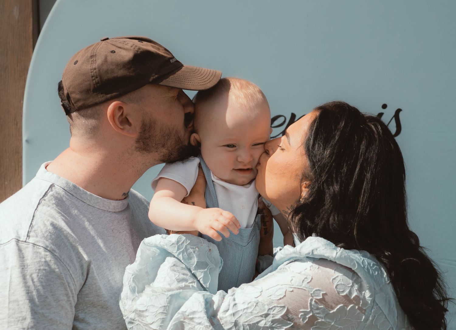 Couple kissing their smiling baby on the cheeks outdoors against a blue wall.