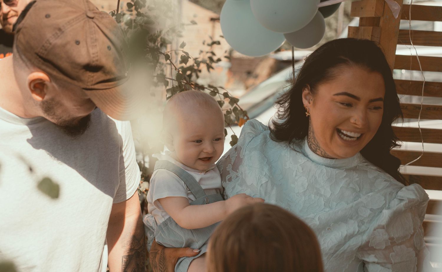 Young couple laughing with happy baby at outdoor celebration with pastel balloon decorations.