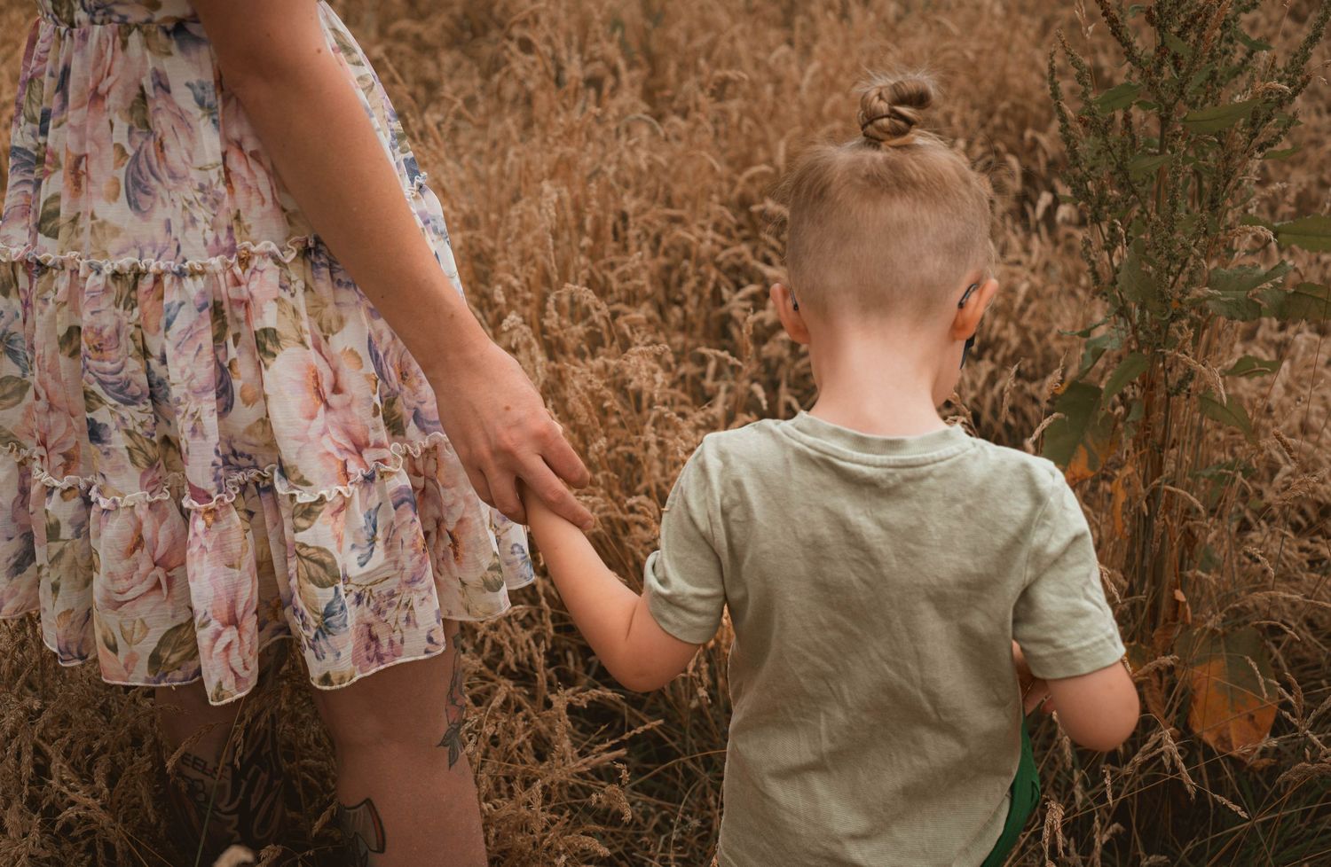 A young child holds hands with an adult in a floral dress while walking through a golden field.