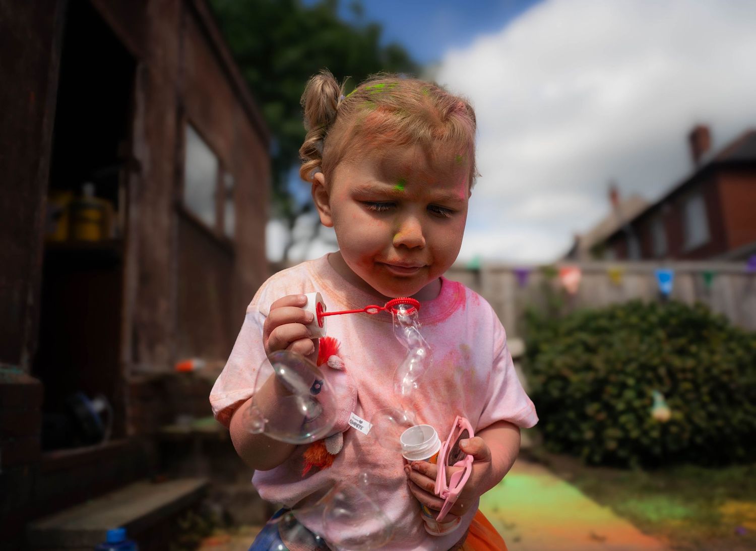 Young toddler with green face paint holding a toy outdoors during a colorful children's party.