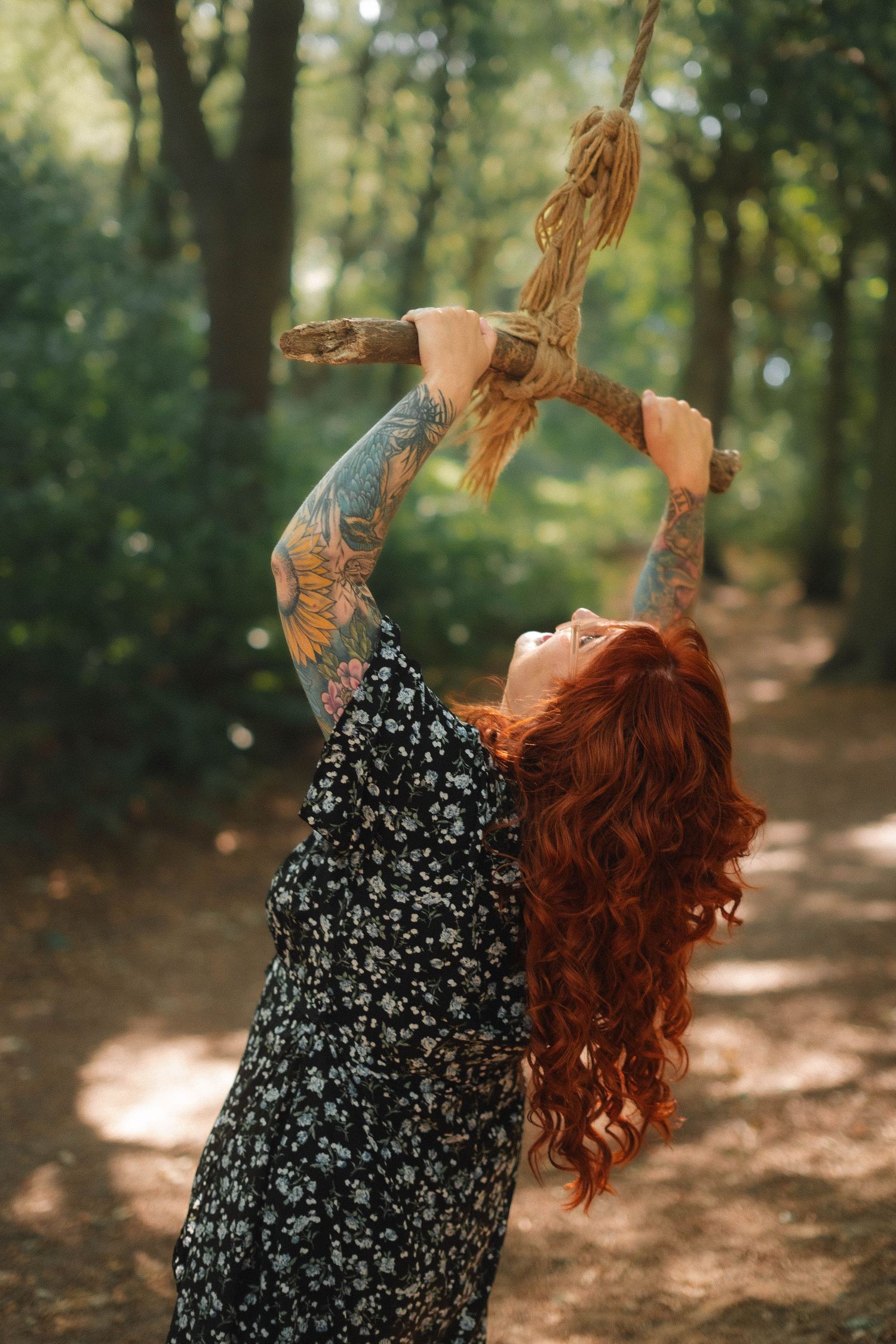 A red-haired tattooed woman reaches up to grab a rope swing in a sunlit green forest.