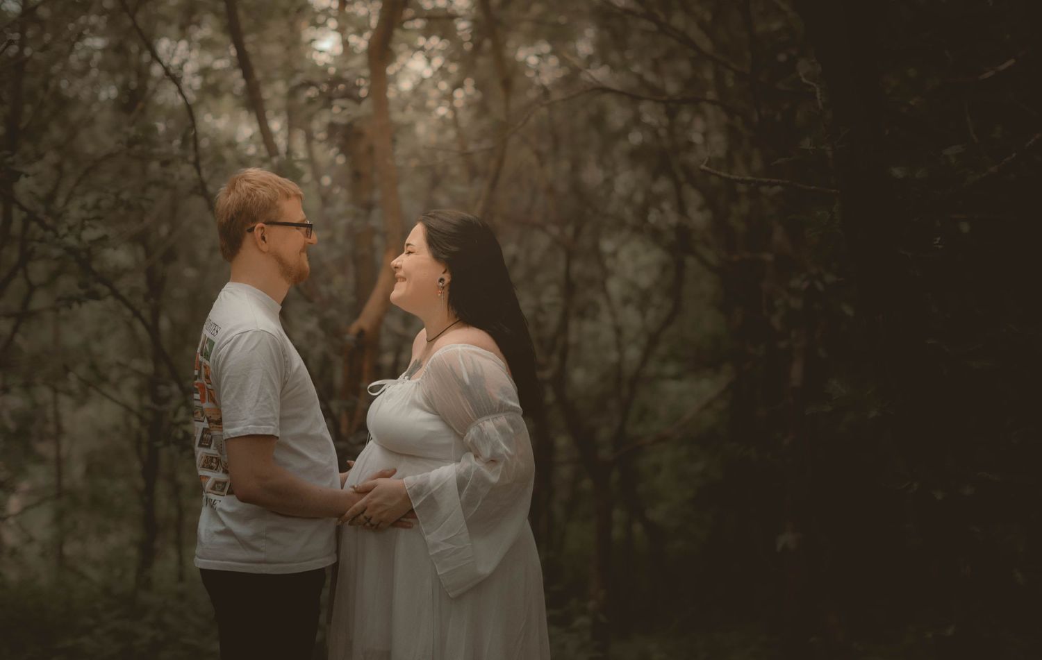 A couple shares a tender moment in a misty forest, the woman in a flowing white maternity gown holding her baby bump.