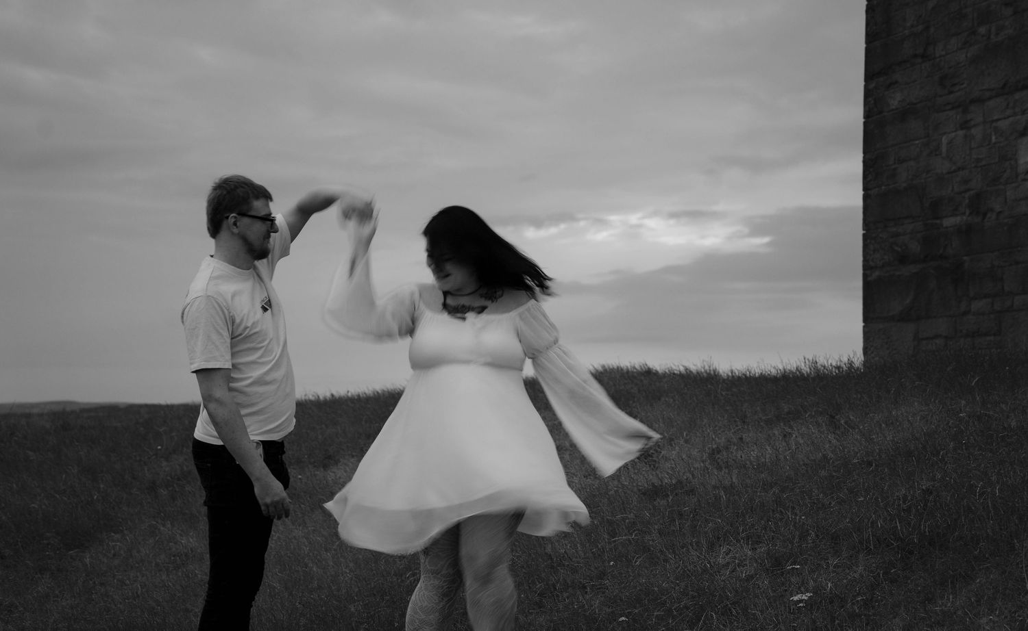 Black and white photo of couple dancing outdoors near a stone structure on a windy day.