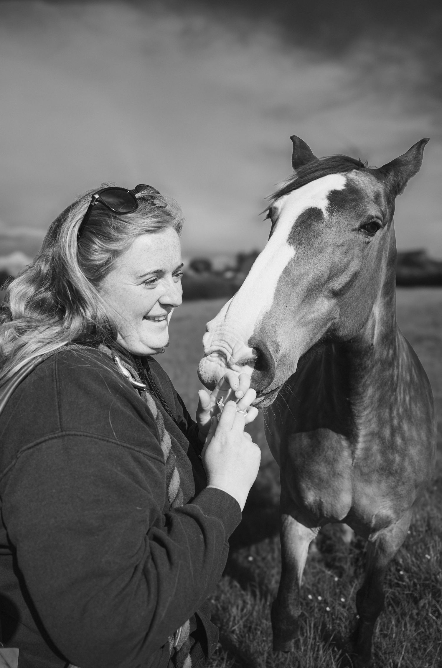 Laughing woman feeding a bay horse with white blaze in black and white photo.