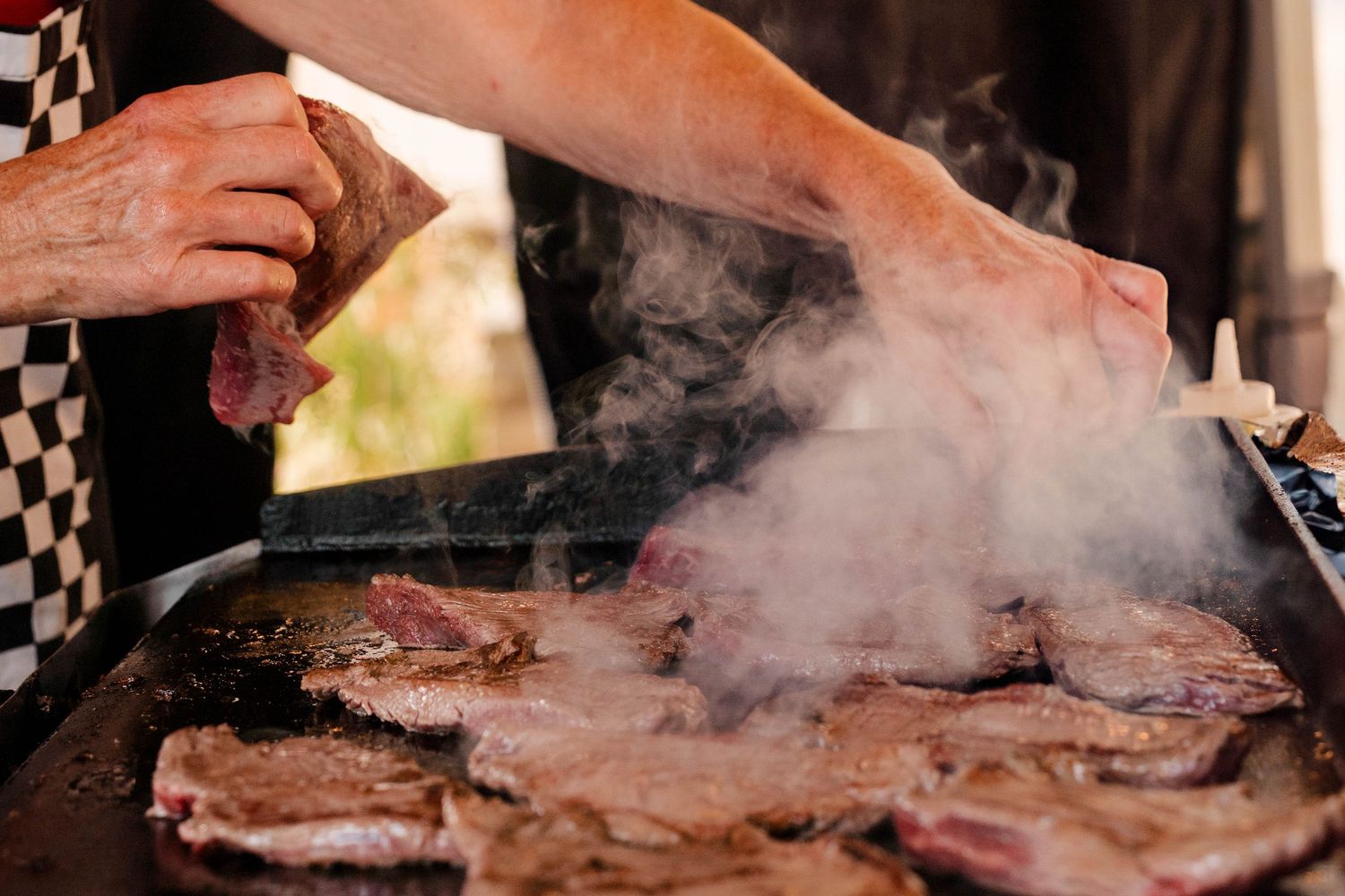 Chef grilling steaks on a large flat top griddle with steam rising at an outdoor event.