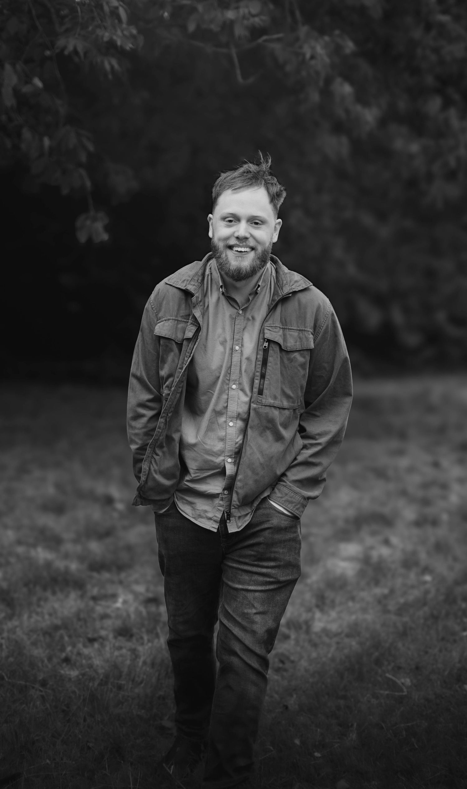 Smiling bearded man in jacket stands in a grassy field in a black and white portrait photo.