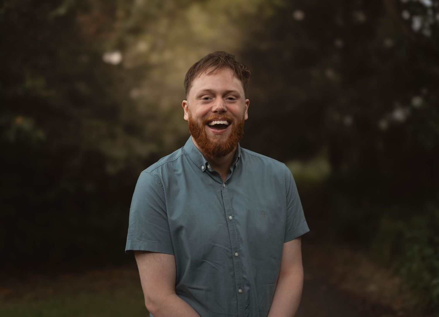Happy red-bearded man laughing outdoors surrounded by lush green trees in natural light.