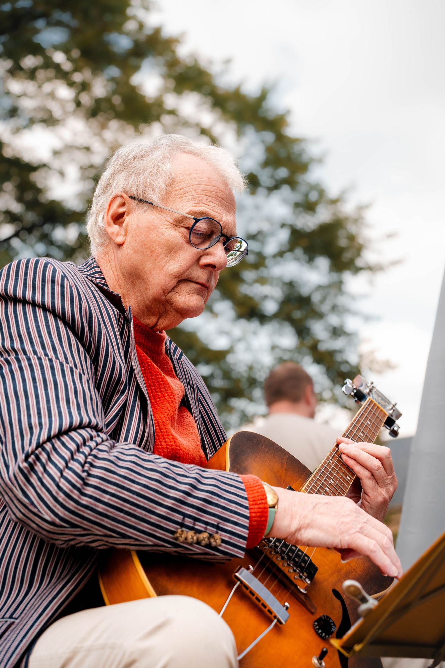 Elderly man playing jazz guitar outdoors wearing striped blazer and red tie.